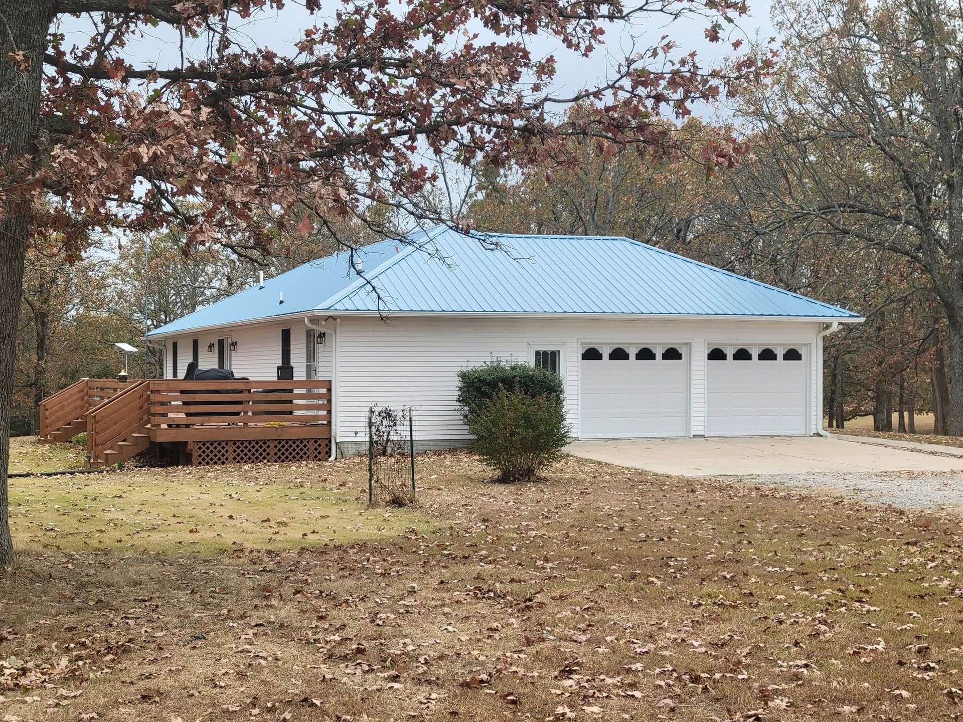 White house with blue metal roof, attached two-car garage, and wooden deck. Surrounded by fall trees.