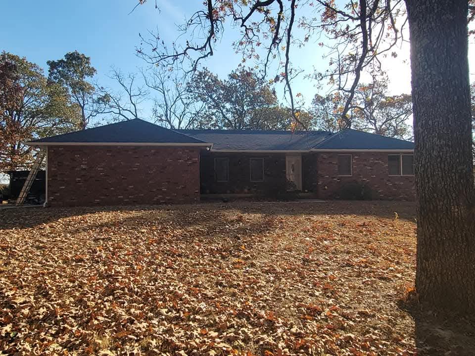Brick ranch-style house with dark roof surrounded by trees and a leaf-covered yard on a sunny day.
