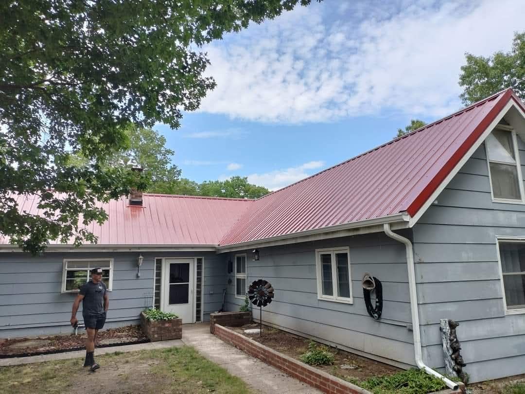 A house with a red metal roof and gray siding. A person is outside, in front.