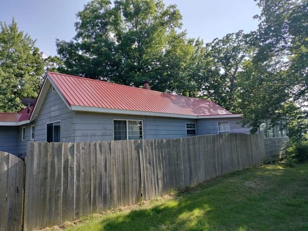 A weathered gray house with a red metal roof behind a wooden fence, surrounded by trees.