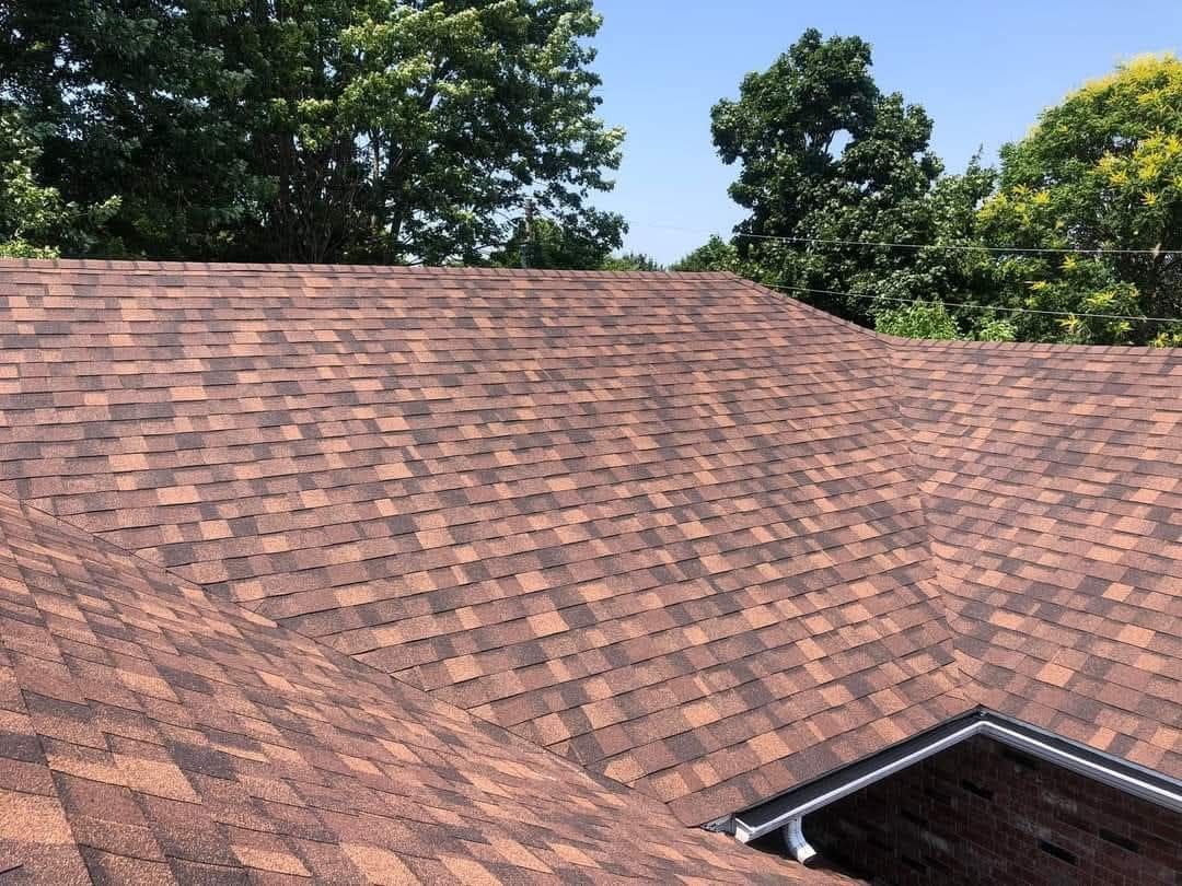 Brown asphalt shingle roof on a house, under a blue sky, with trees in the background.