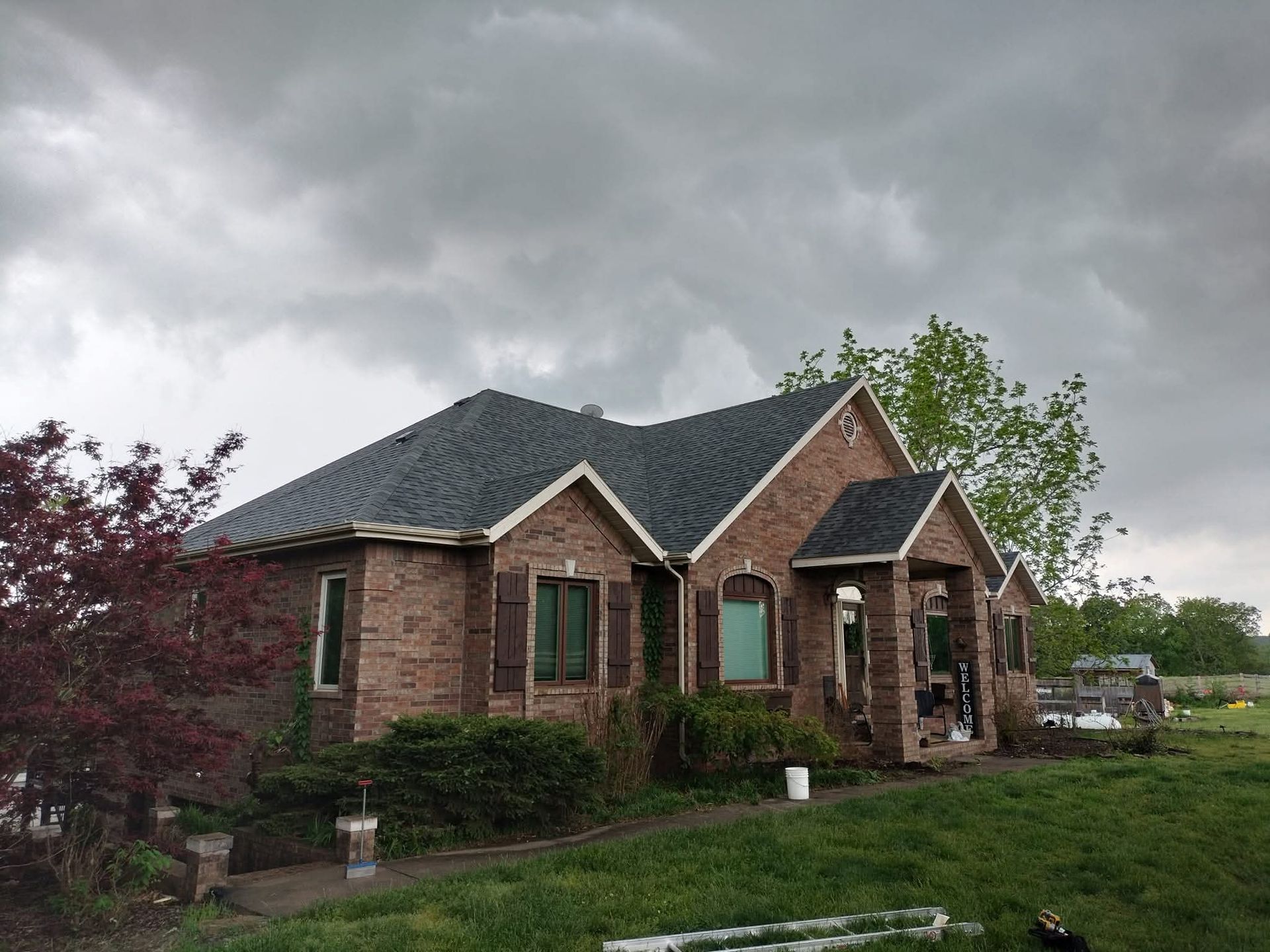 Brick house with dark roof under cloudy sky. Green grass, bushes, and trees surround the home.