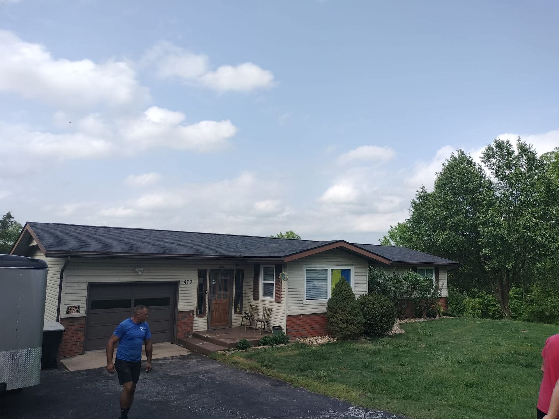 House with new roof and asphalt driveway. Man walks on driveway. Green lawn and trees. Cloudy sky.