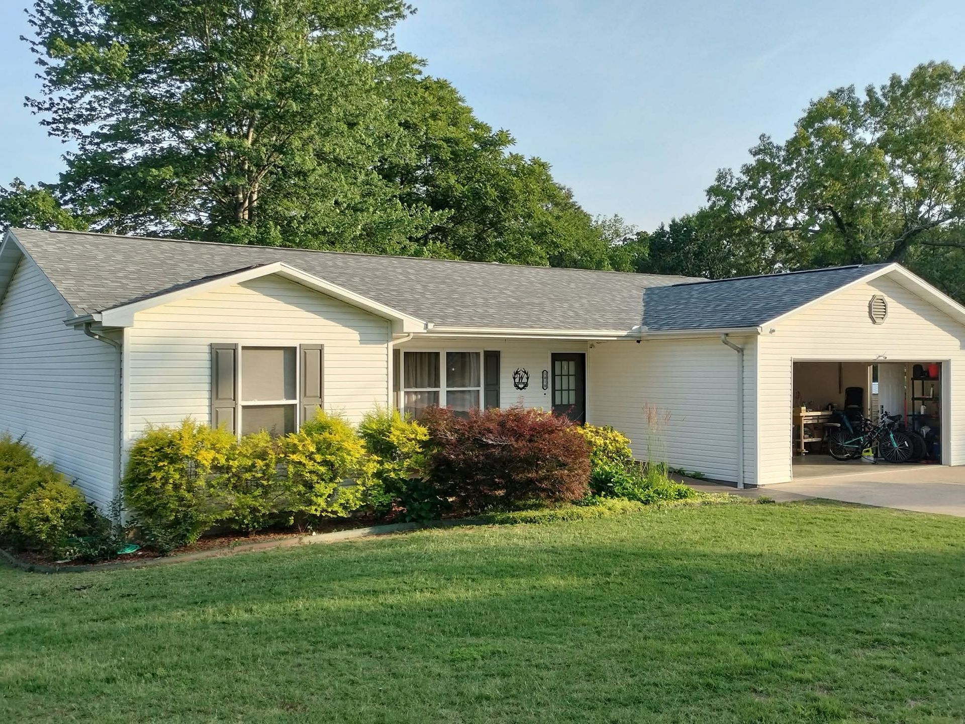 White house with a gray roof, attached garage, and lawn with shrubs and trees.