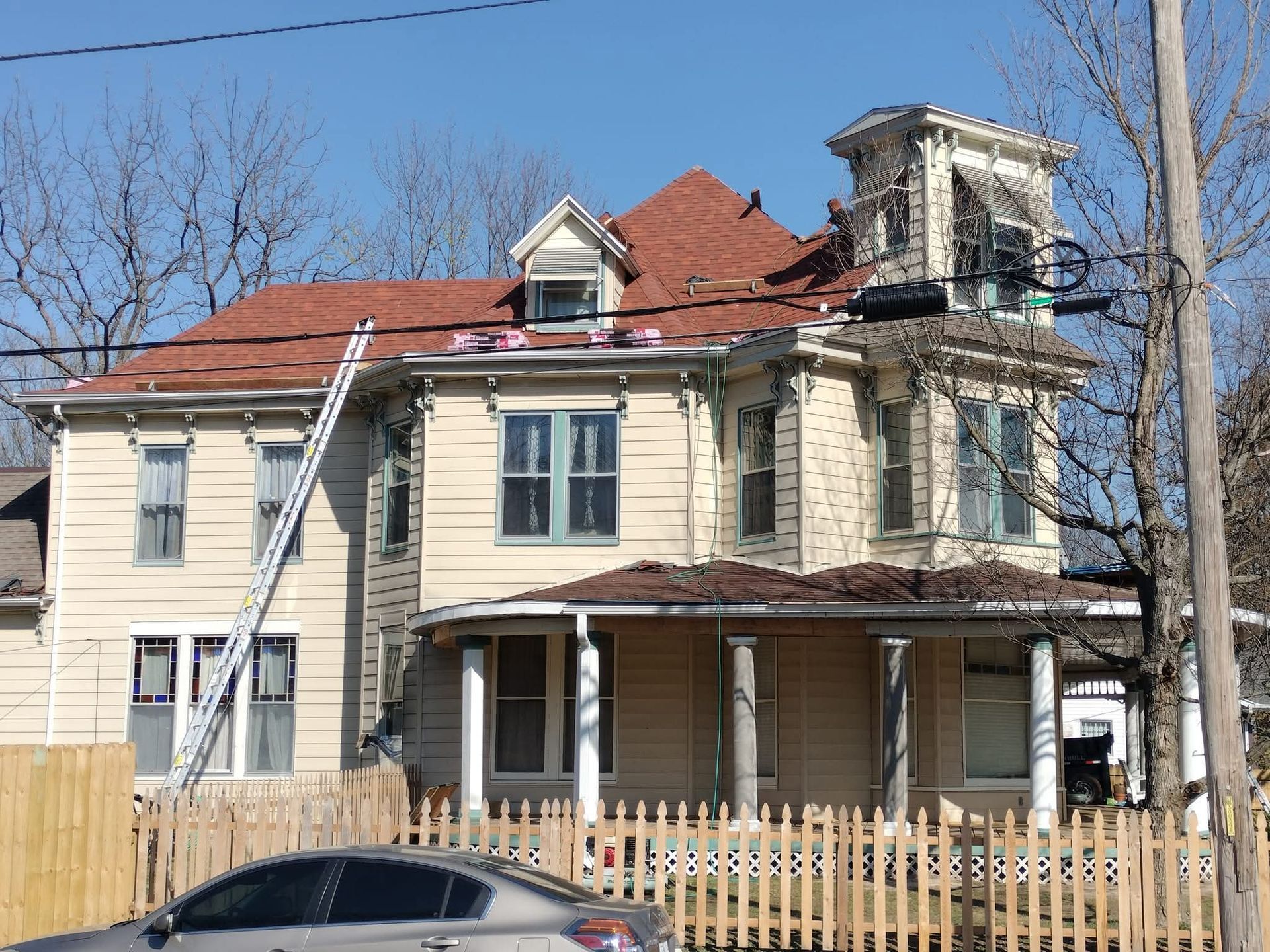 Beige two-story house with red roof, ladder leaning against it, and a wooden fence in front.