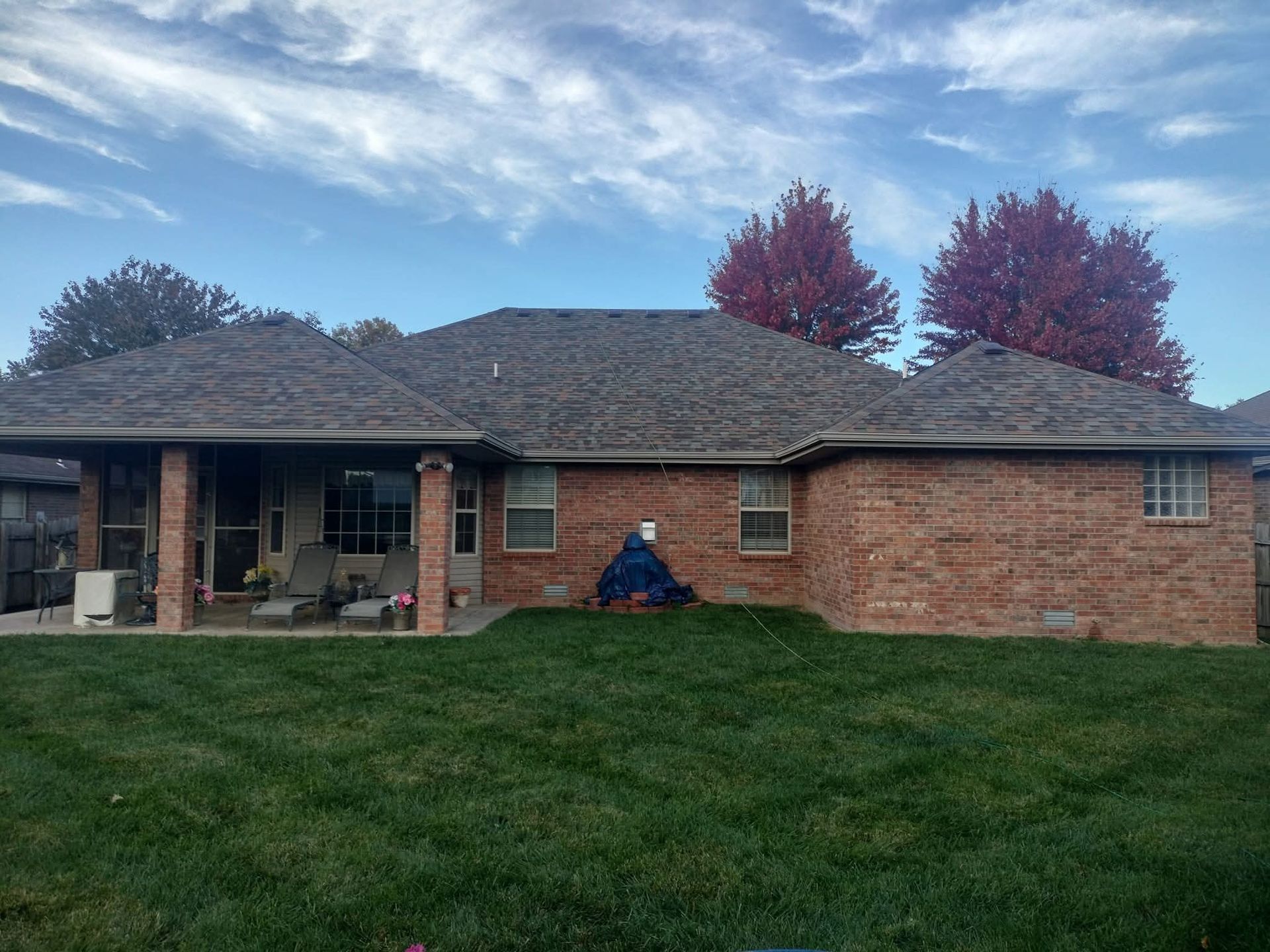 Back of a brick house with a dark roof and a covered patio, set against a lawn and cloudy sky.