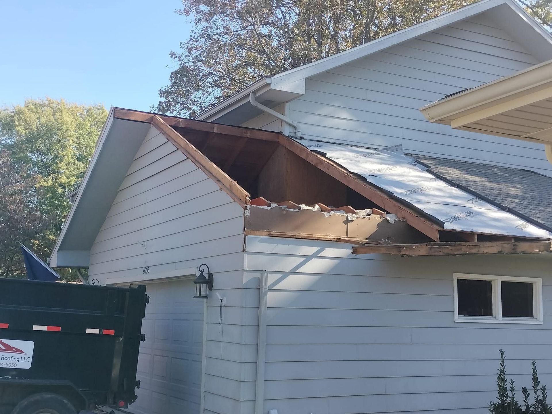 House with roof damage, missing sections exposing wooden beams and underlayment; dumpster parked nearby.