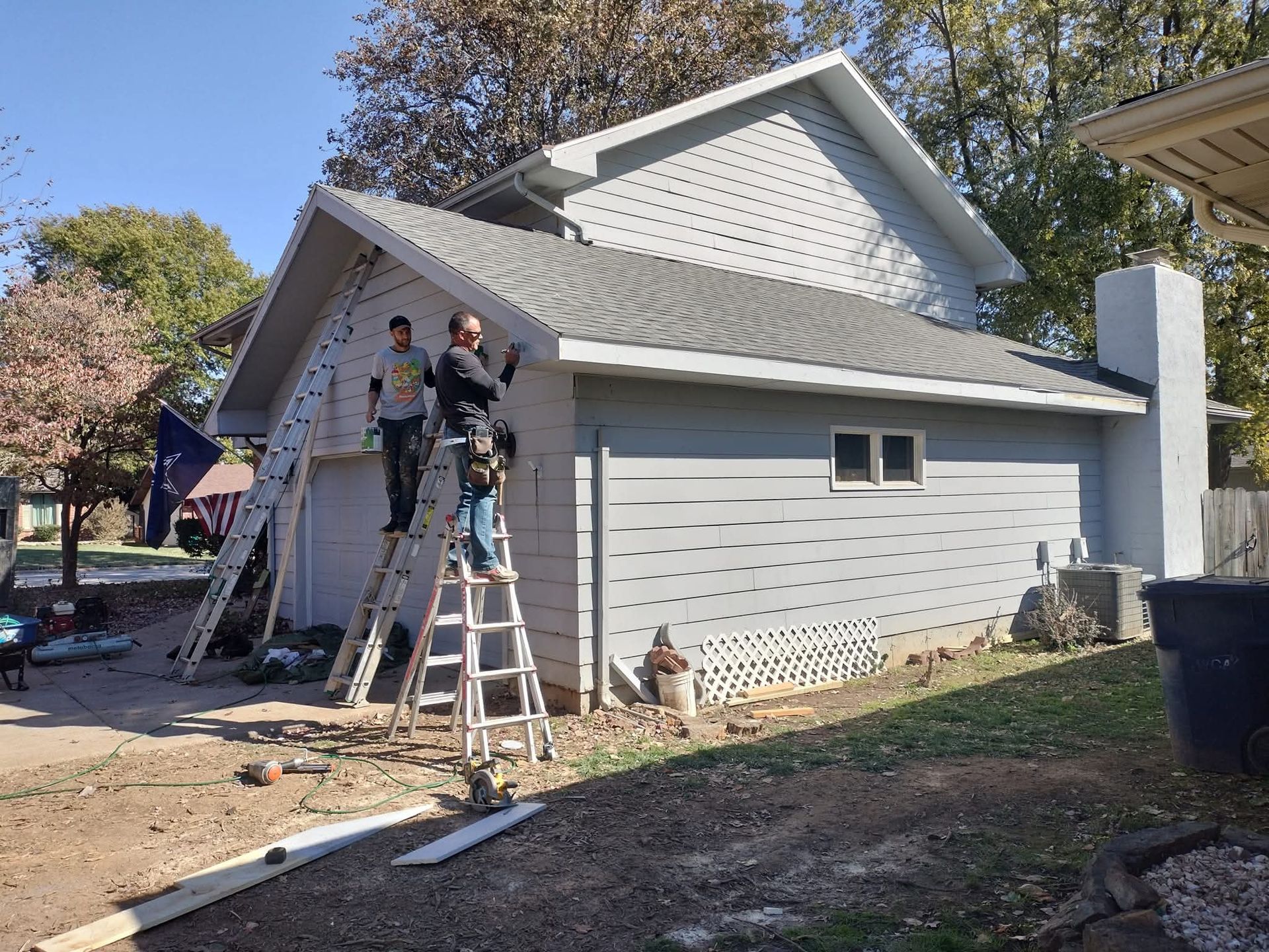 People on ladders siding a gray house with a chimney on a sunny day.