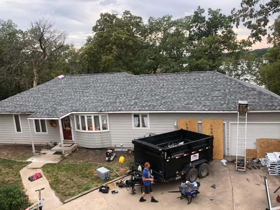 House with new gray roof, construction site in front. Dumpster, worker, ladder. Green trees in background.