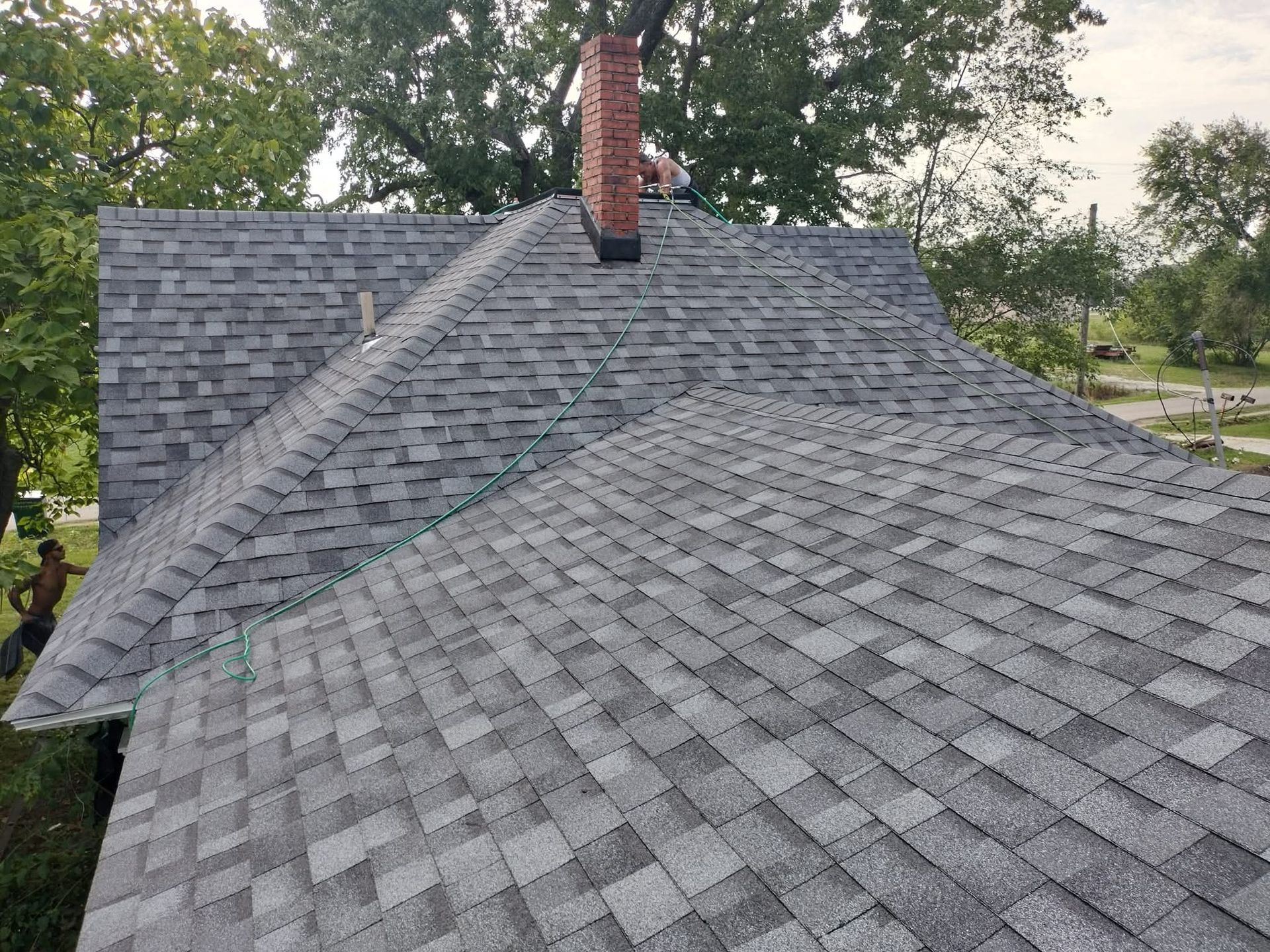 Newly shingled gray roof with a brick chimney; person working near the edge.