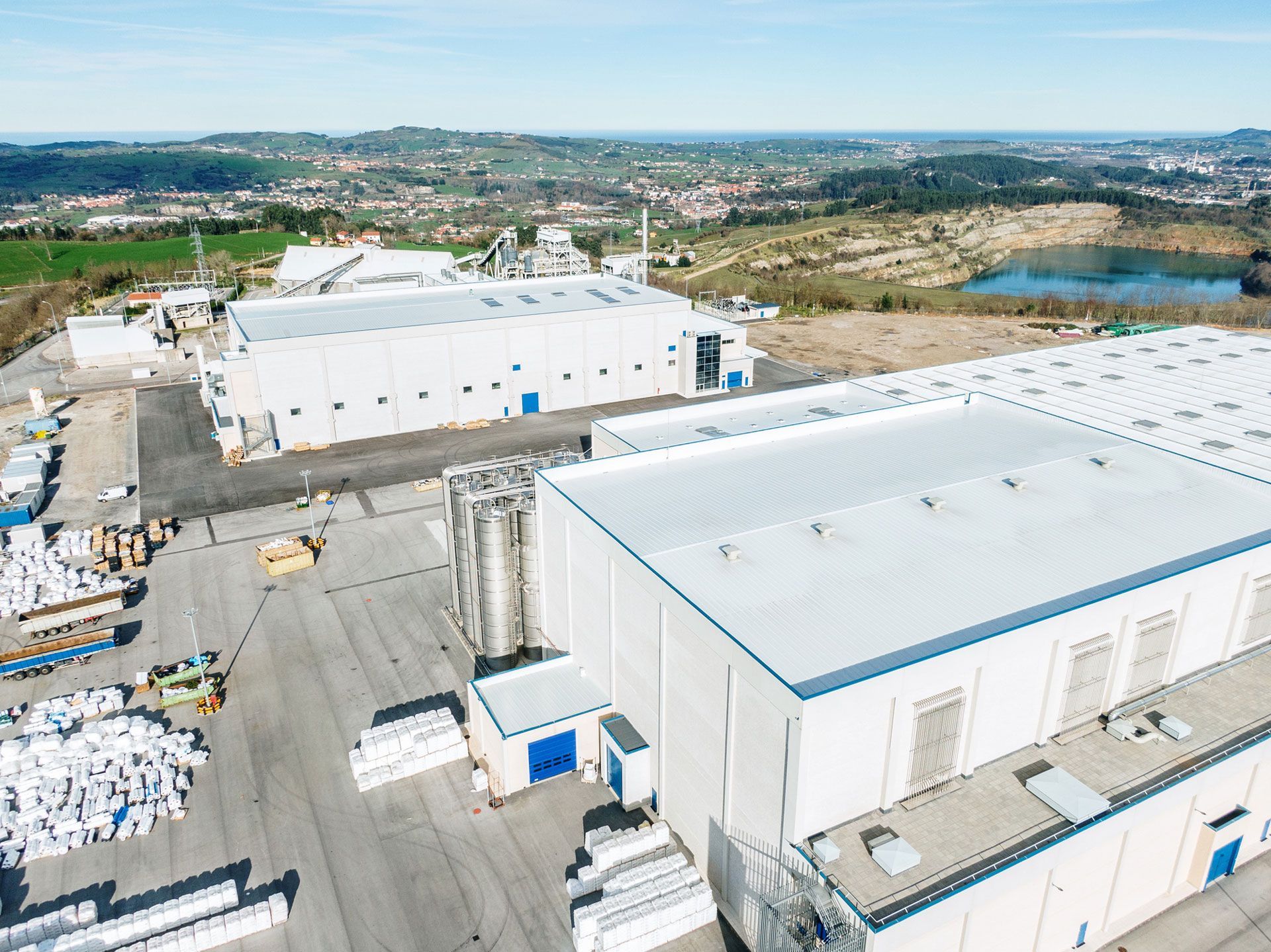 Aerial view of an industrial complex with multiple white buildings, a paved lot, and a distant landscape with a lake. Aerial view of an industrial complex with multiple white buildings, a paved lot, and a distant landscape with a lake.