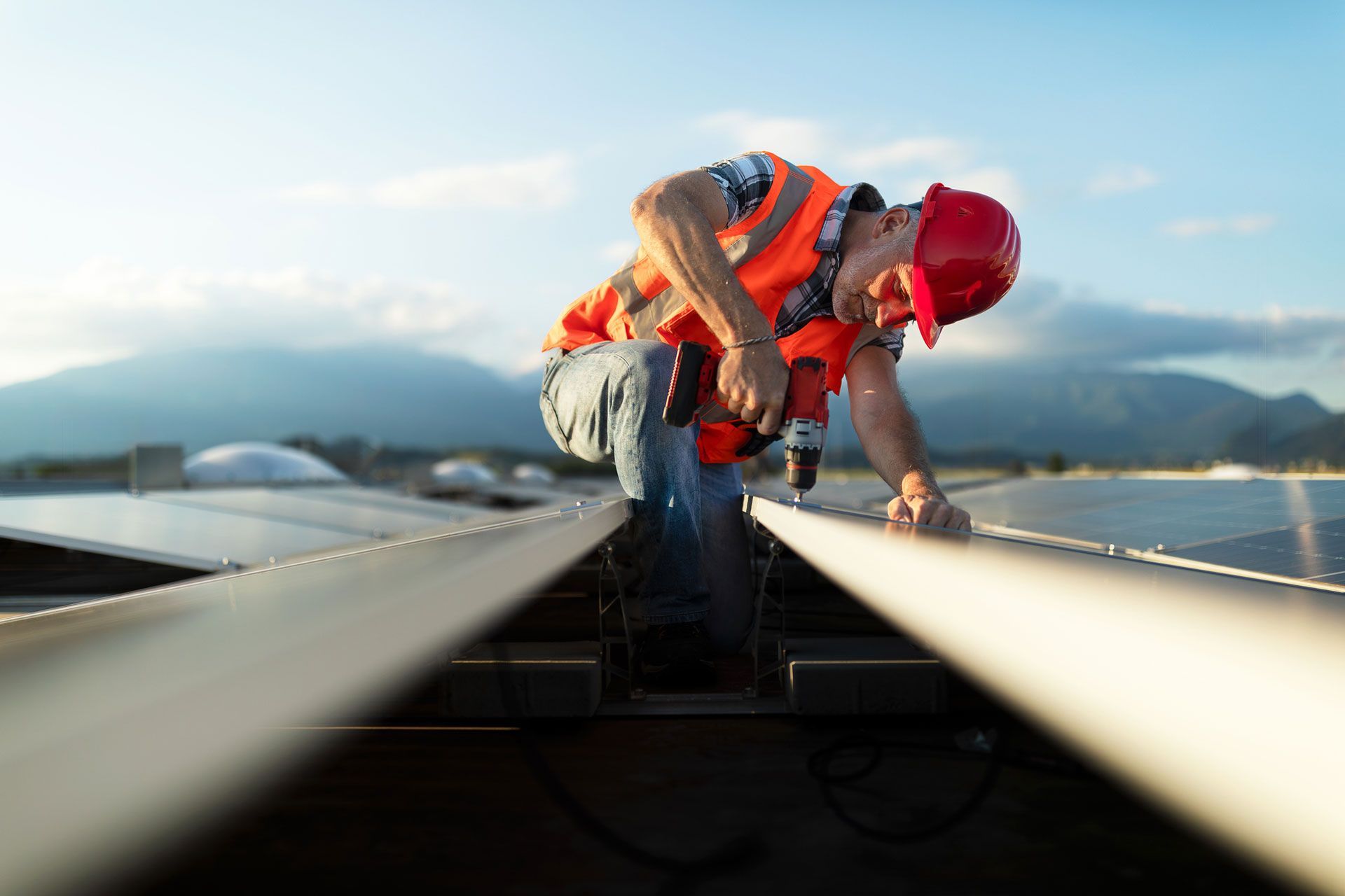 Construction worker in safety vest and hard hat using a drill on rooftop solar panels. Mountains in background.