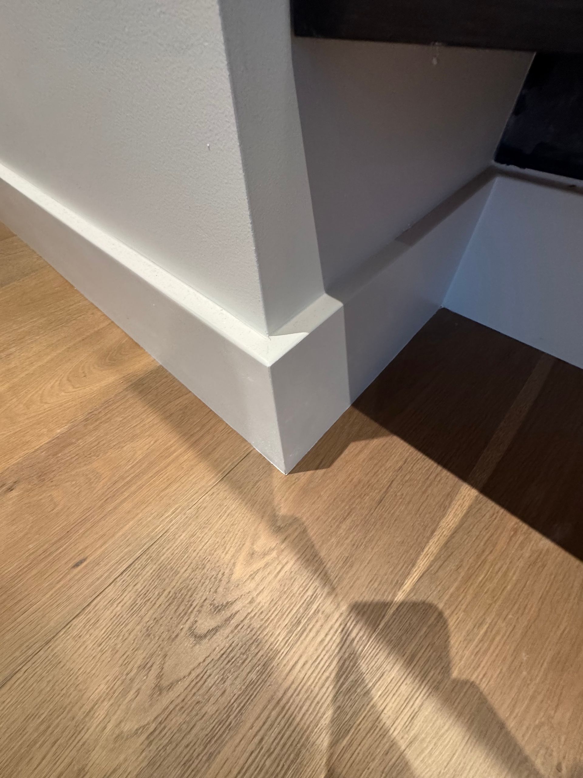 White wall corner with baseboard beside a light wood floor and stair opening shadows