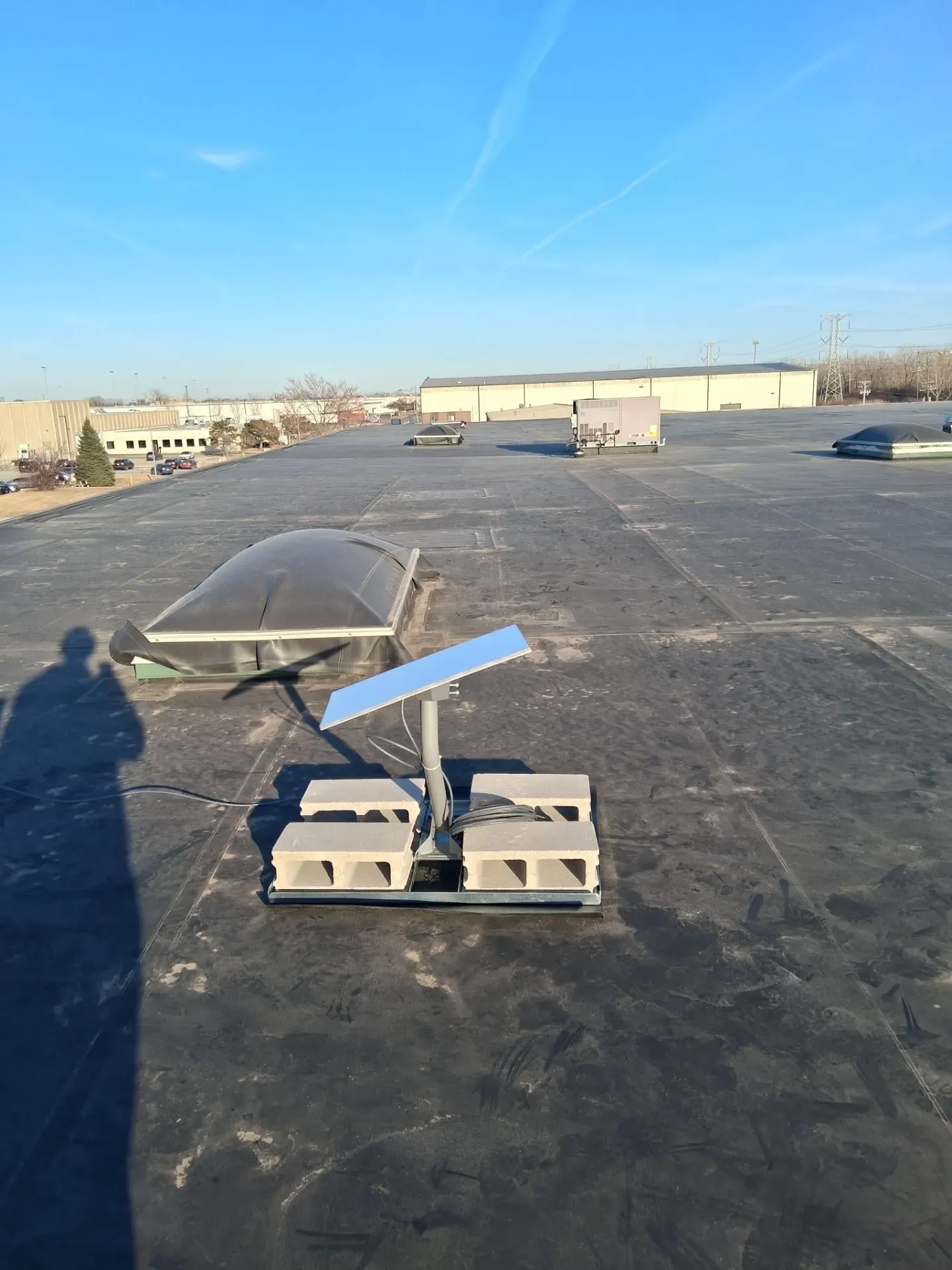 A solar panel mounted on a weighted concrete base sits on a flat, dark commercial roof under a clear blue sky.