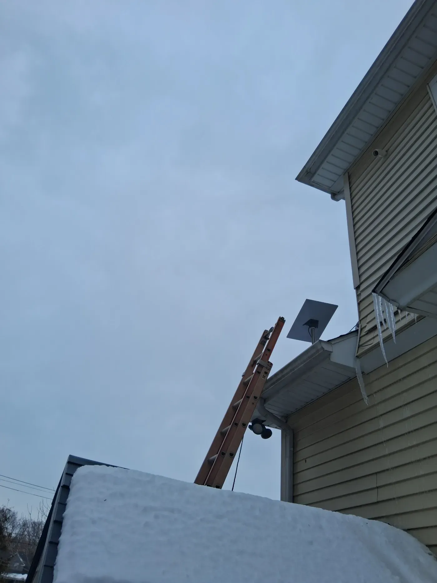 A wooden ladder leans against the side of a beige house near a snowy roof, with icicles hanging from the eaves.