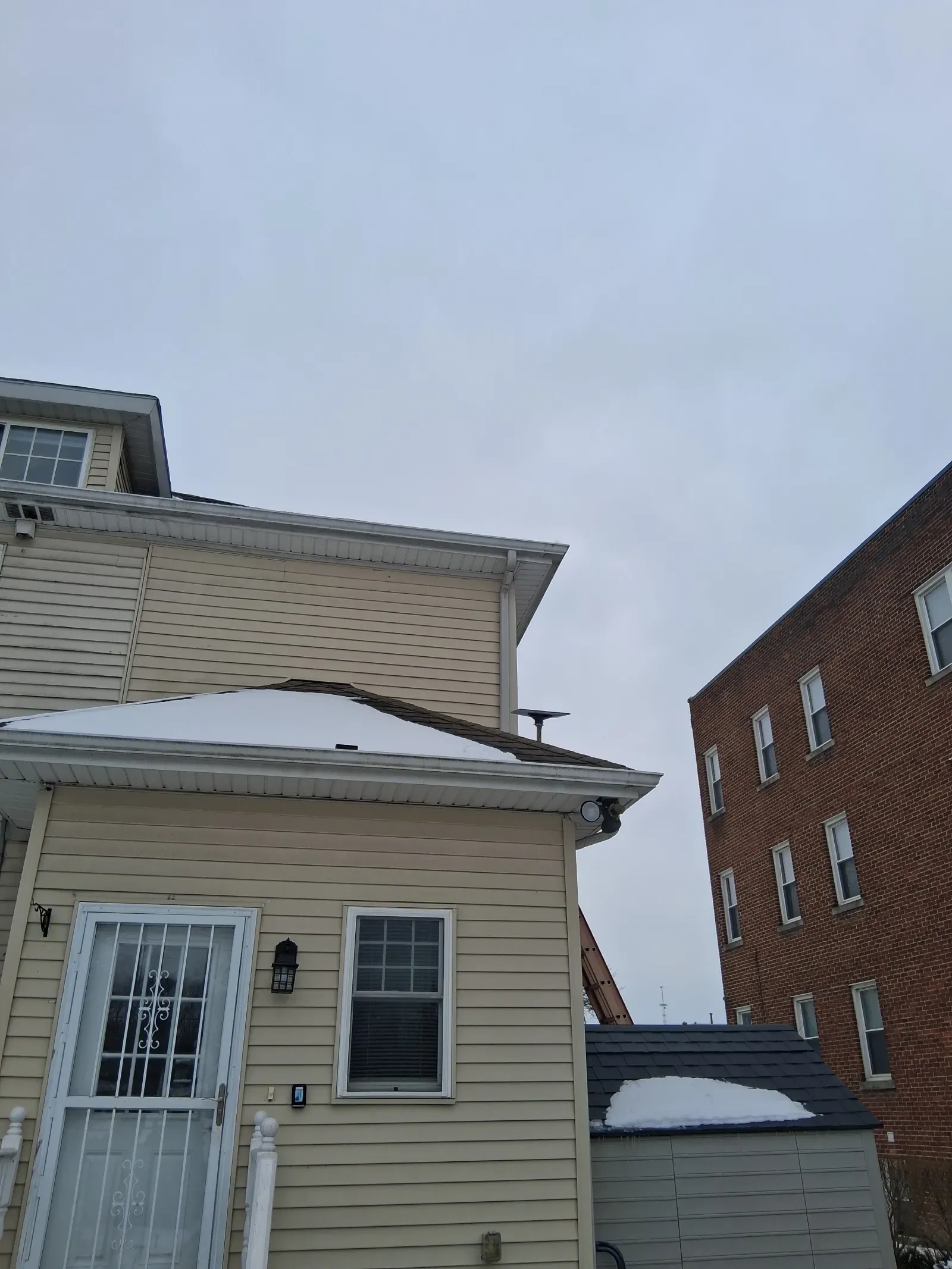 Beige home with vinyl siding and a snowy roof, viewed from an angle against a cloudy sky next to a brick building.