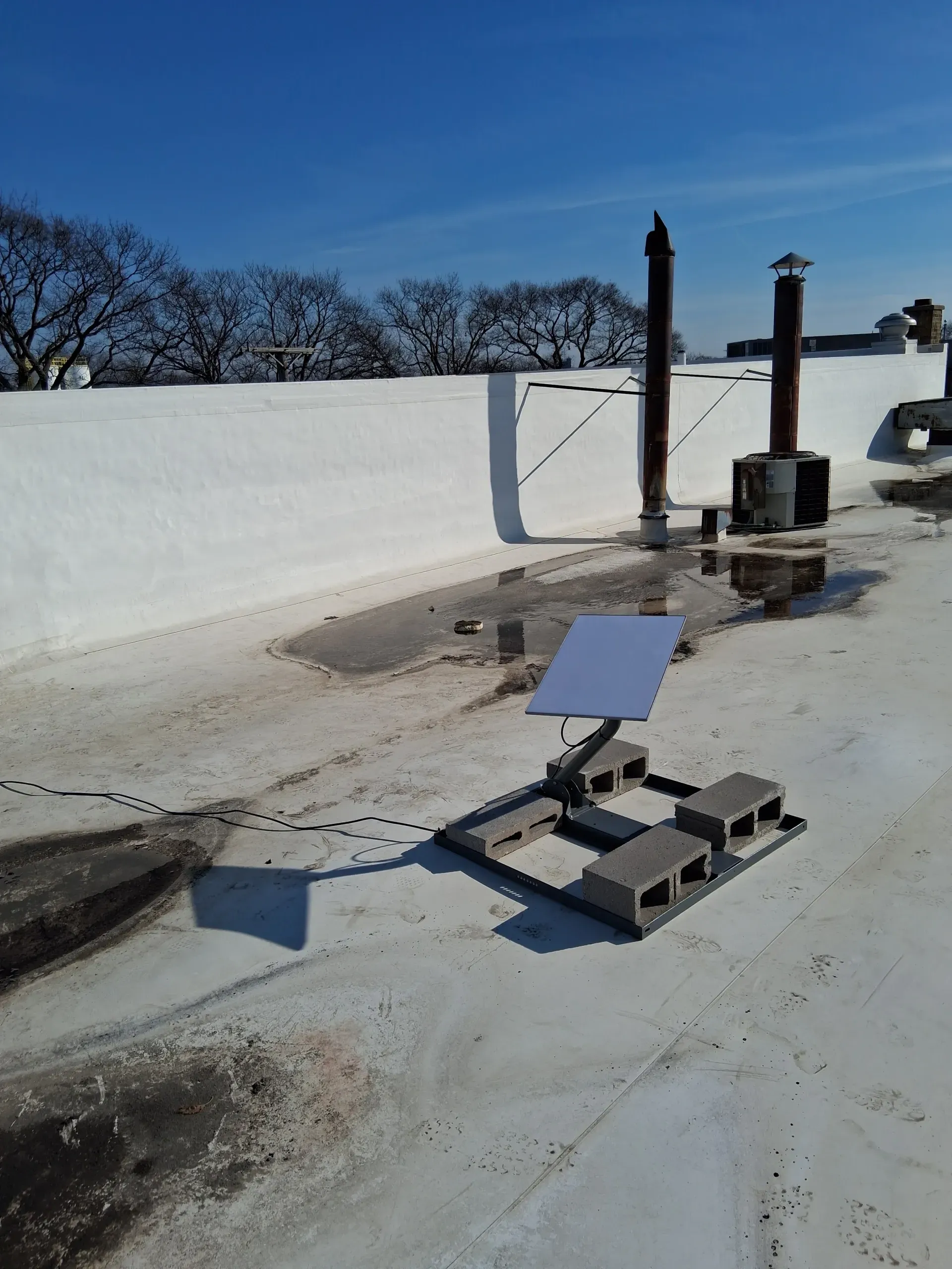 A Starlink satellite dish mounted on concrete blocks sits on a flat white rooftop under a clear blue sky.