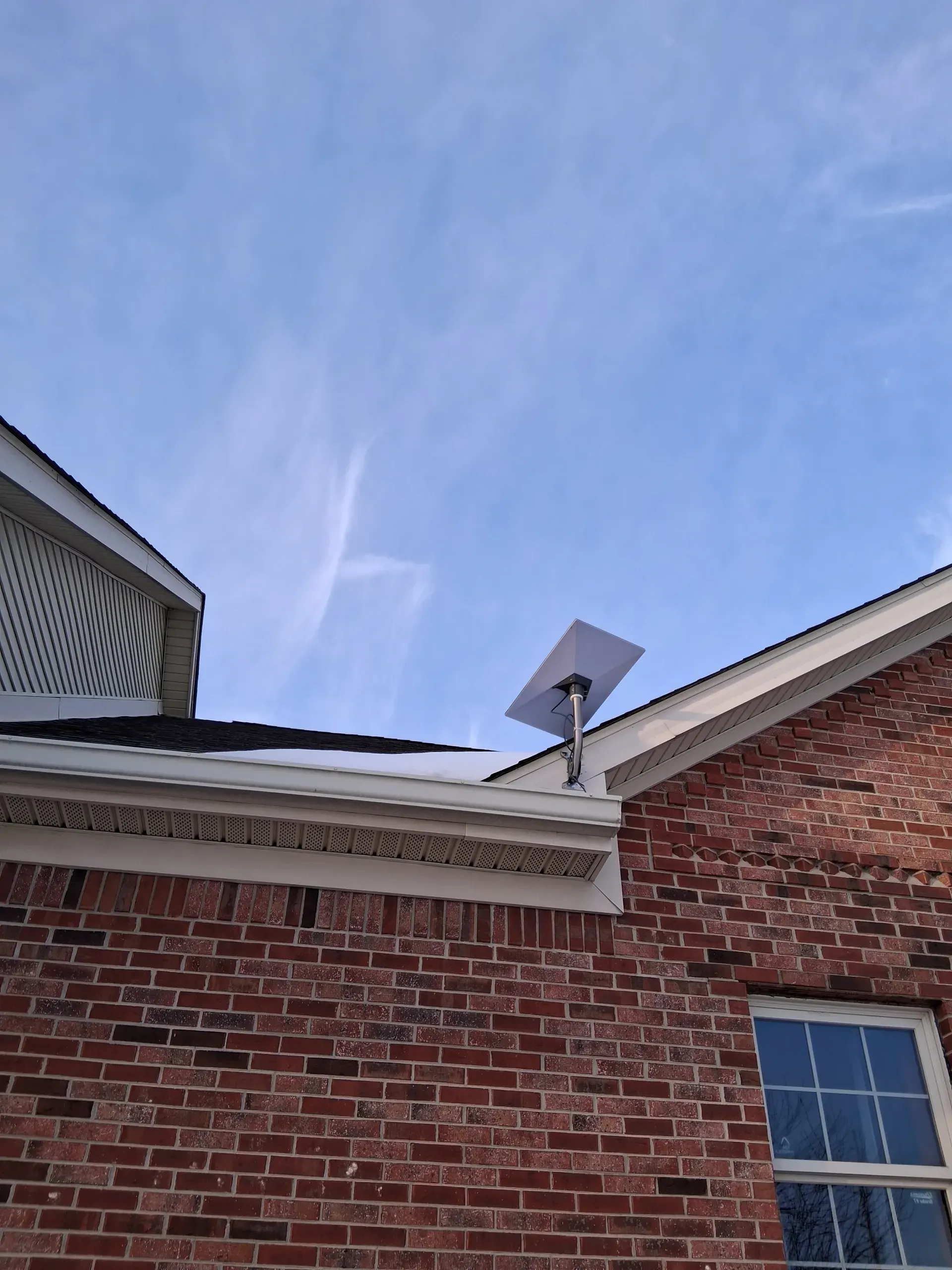 A Starlink satellite dish mounted on the roof of a red brick house against a blue, partly cloudy sky.