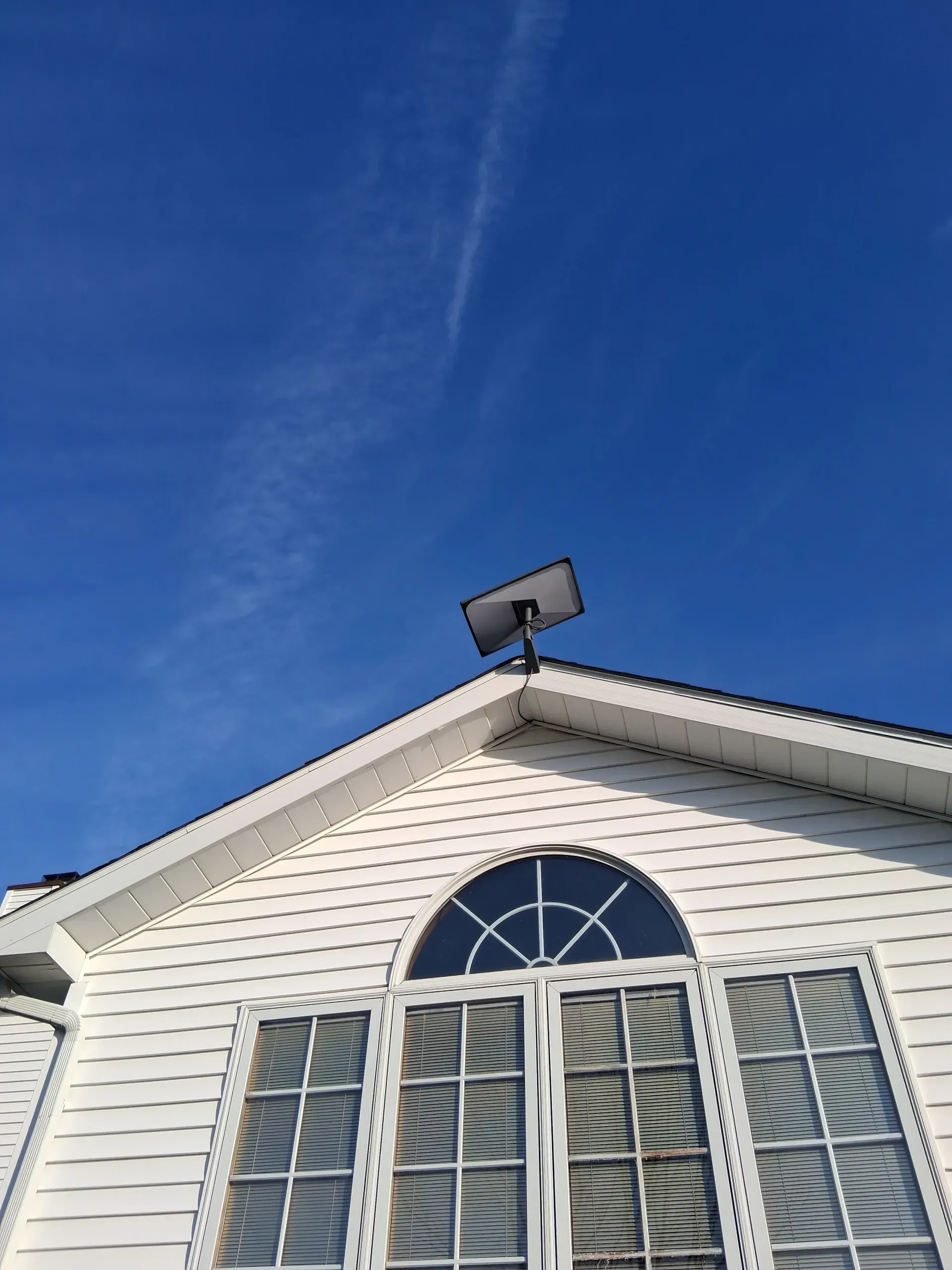 A Starlink satellite dish mounted on the peak of a white vinyl-sided house against a bright blue sky.