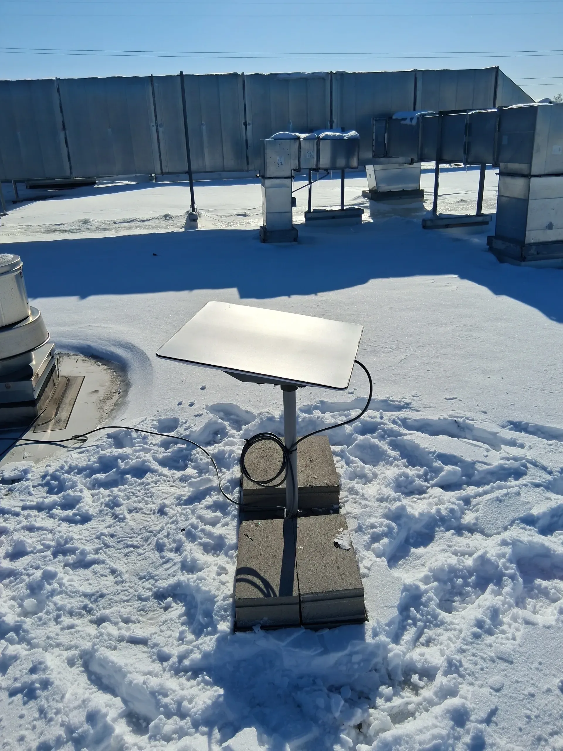 A satellite dish mounted on a stand with concrete blocks on a snow-covered rooftop with HVAC equipment in the background.