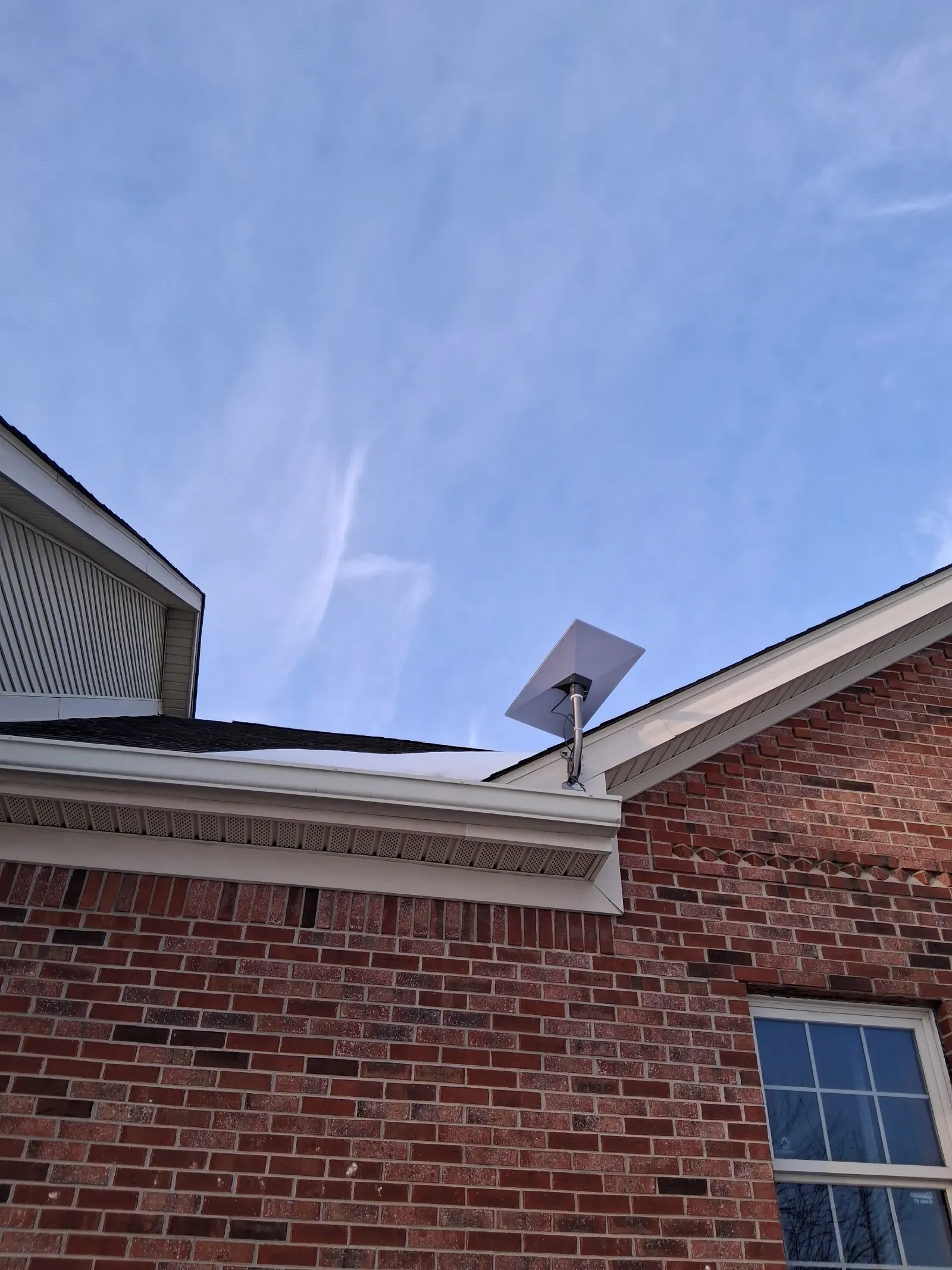 A rectangular satellite internet dish mounted on the roof of a red brick house against a clear blue sky.