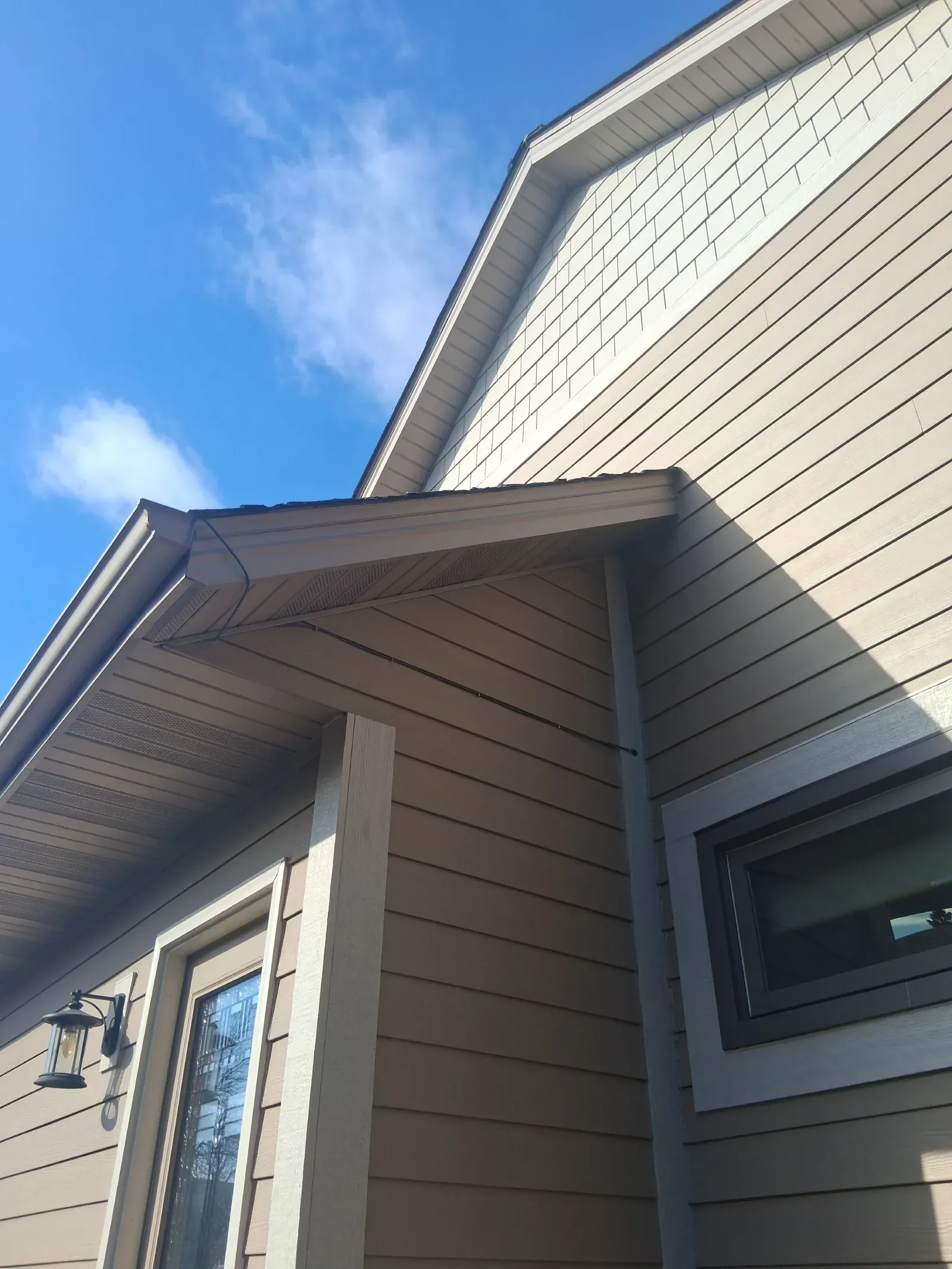 A low-angle view of a beige house exterior with tan horizontal siding, a gabled roof, and a clear blue sky above.