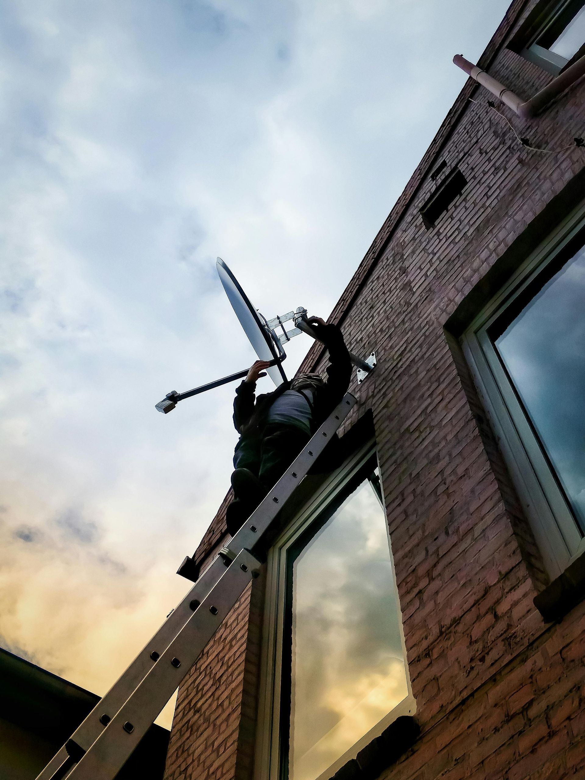 Person on a ladder installing a satellite dish on a brick building, under a cloudy sky.