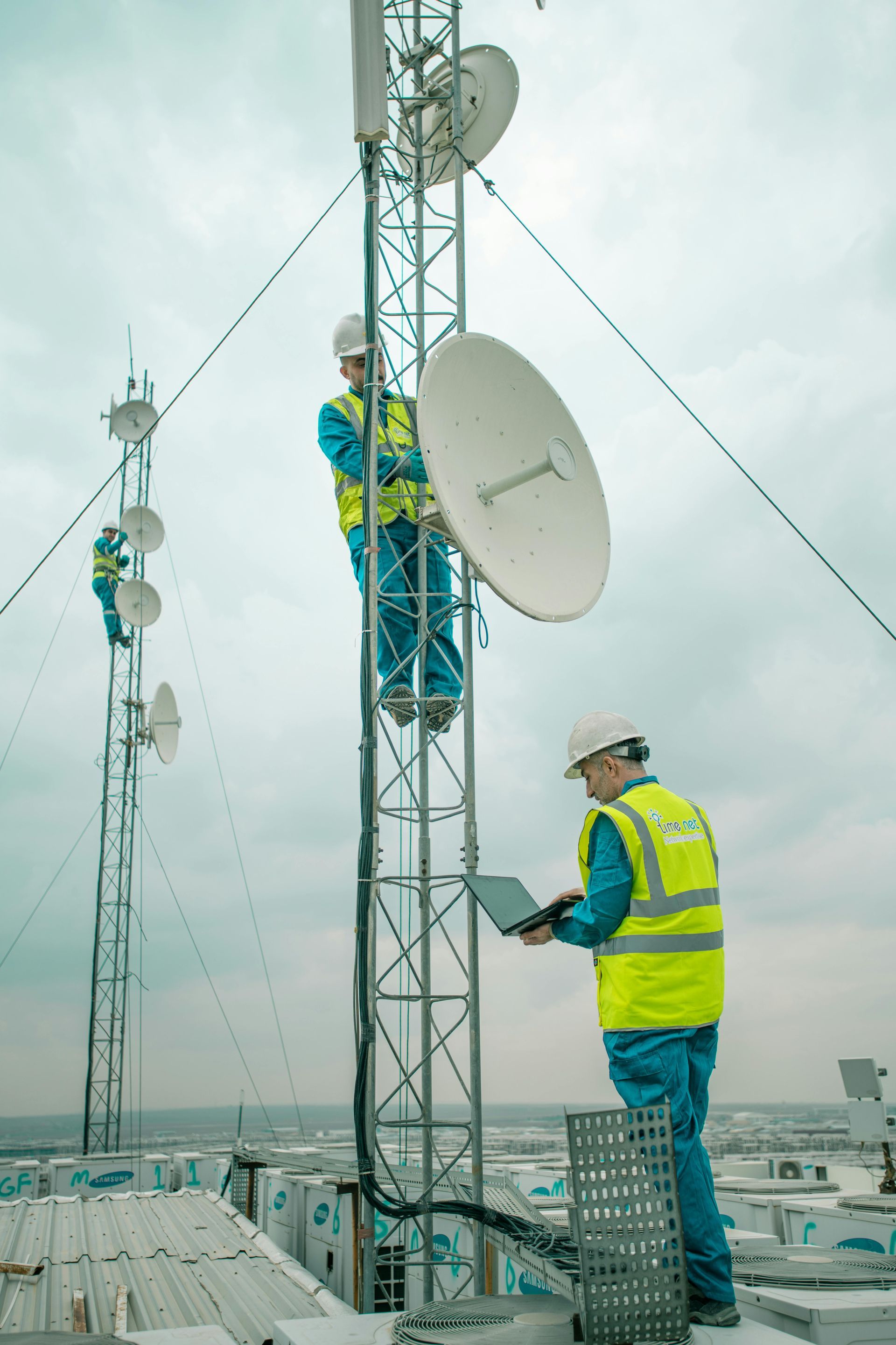 Technicians in safety vests work on a communications tower, one using a laptop while others work above.