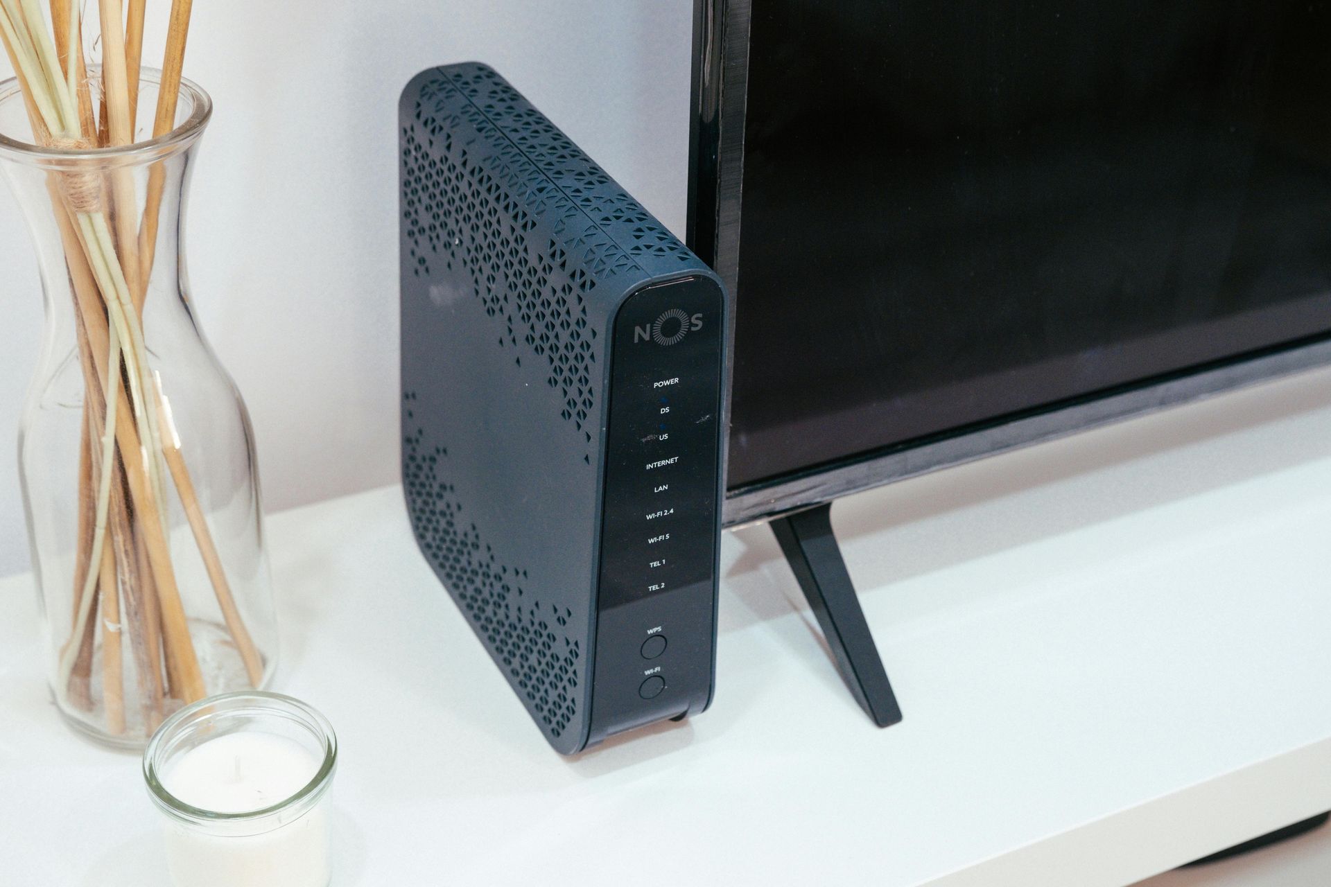 Black modem next to a television on a white shelf, with decorative vase and candle.