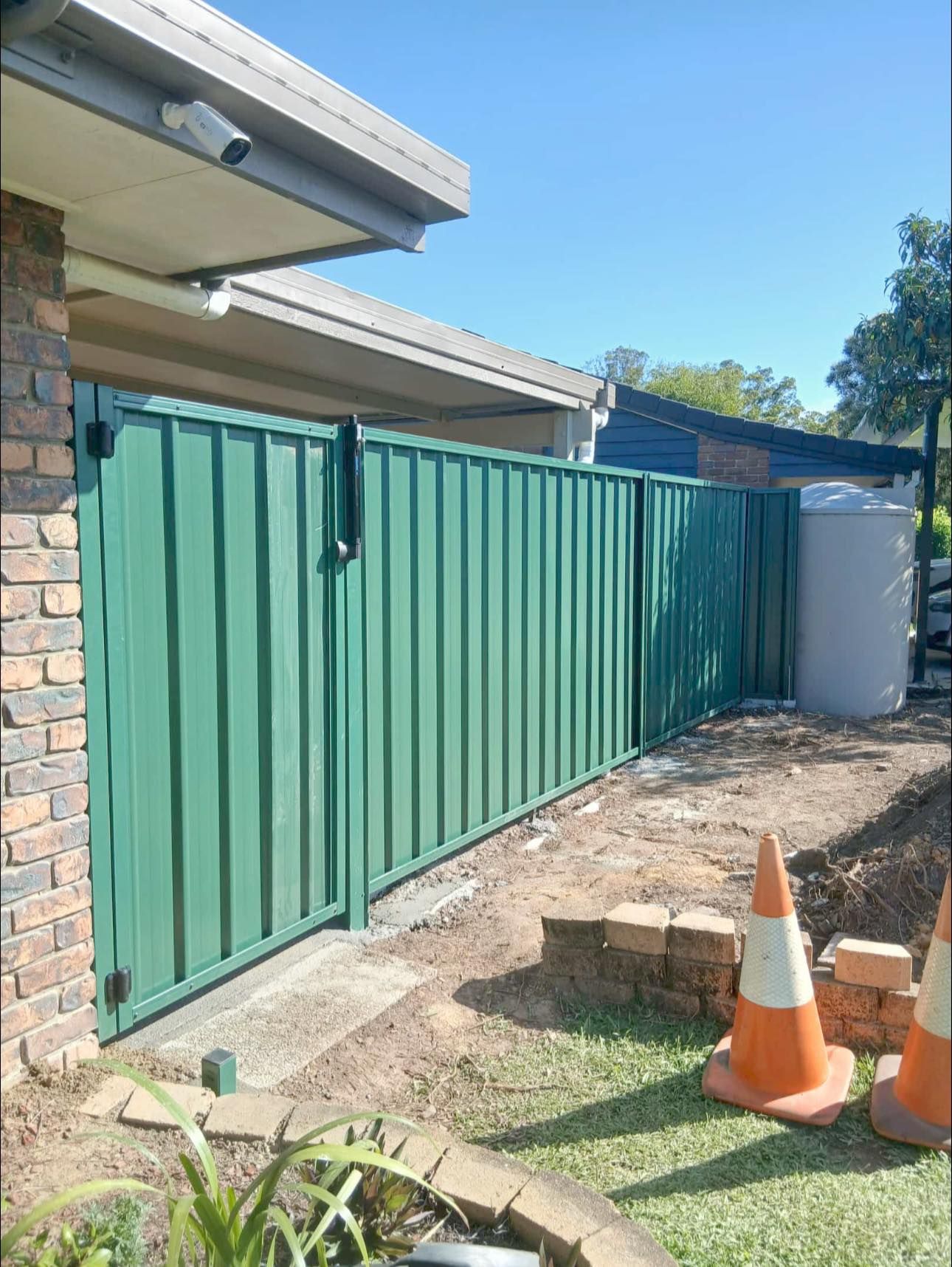 Green metal fence beside a brick building with a water tank and traffic cones — Chore Lovers in Caboolture, QLD