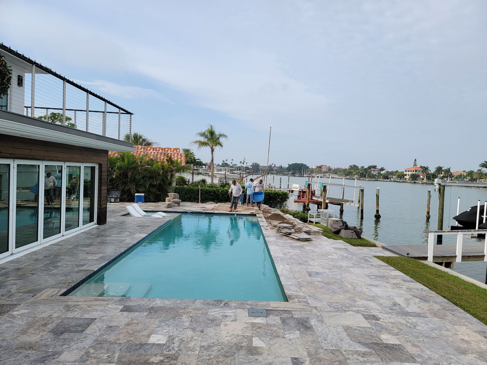 Waterfront pool beside a house, with lounge chairs and people by the dock at sunset