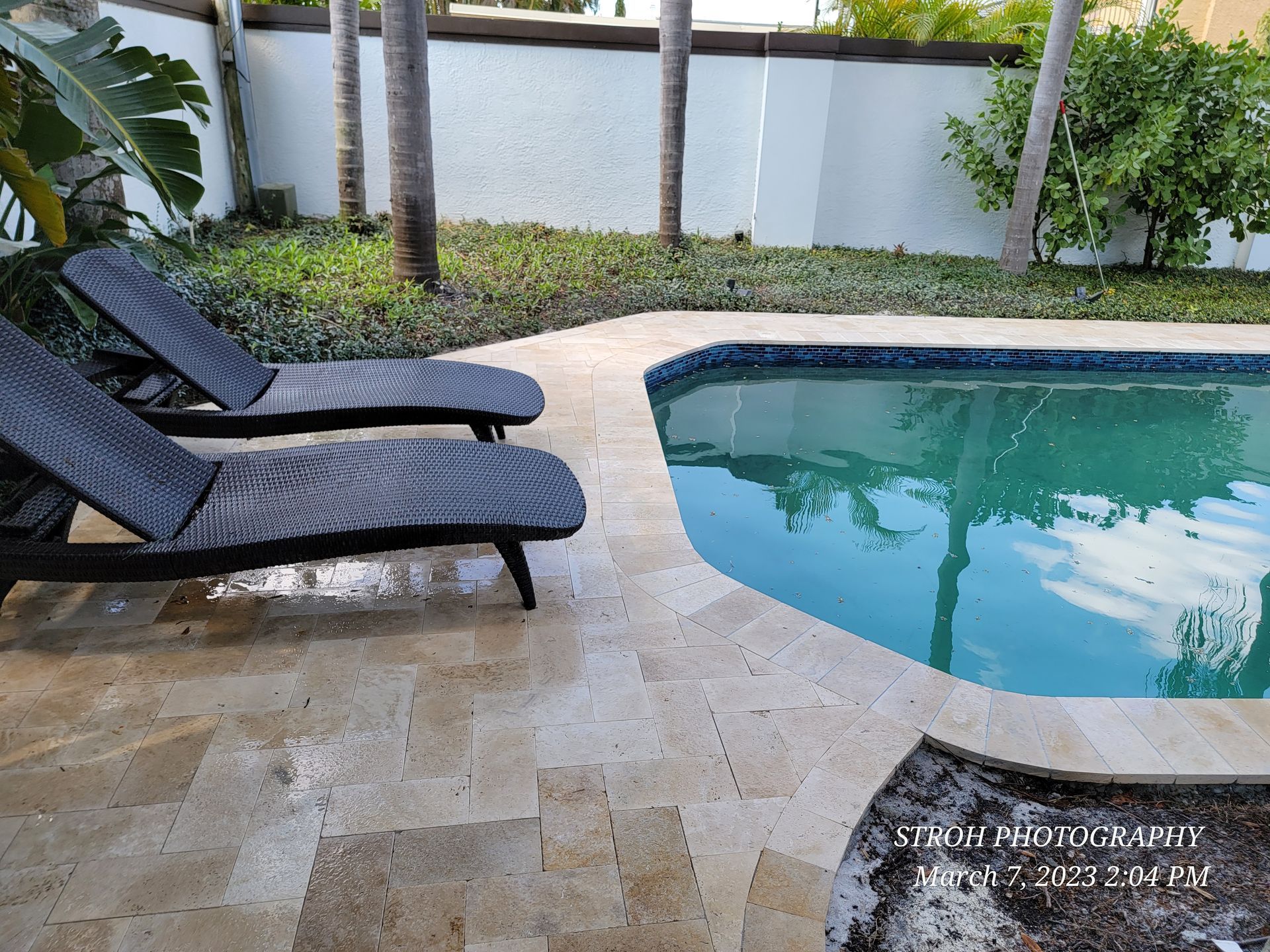 Poolside patio with two black lounge chairs beside a blue swimming pool and white fence.