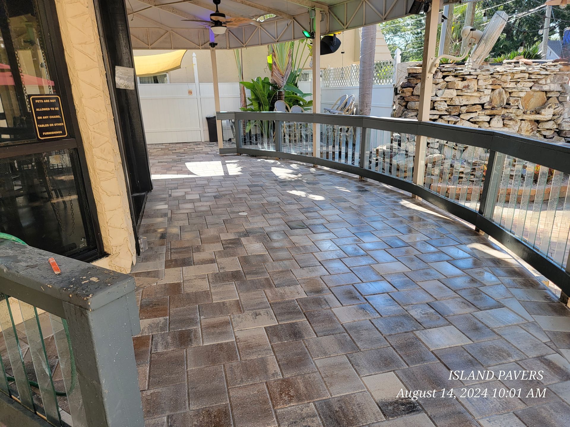 Covered outdoor patio with tiled floor, railing, ceiling fan, and stone wall in bright daylight
