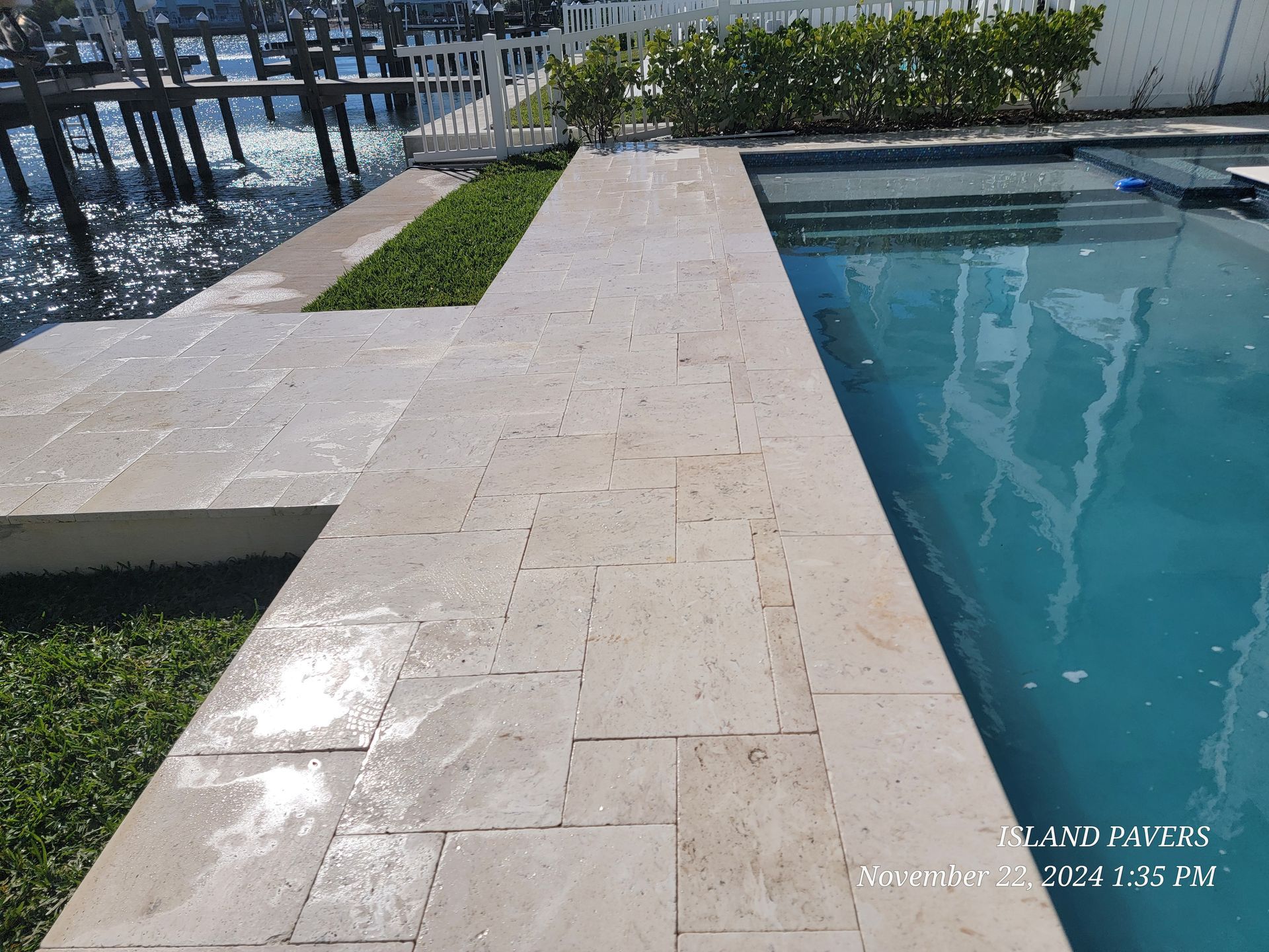 Stone pool deck beside clear blue water with grass borders and a metal fence in the background