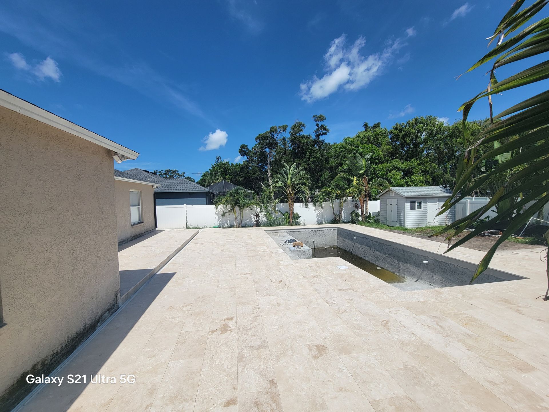 Sunny driveway and patio beside a beige house, with a garage, white walls, and tropical plants.