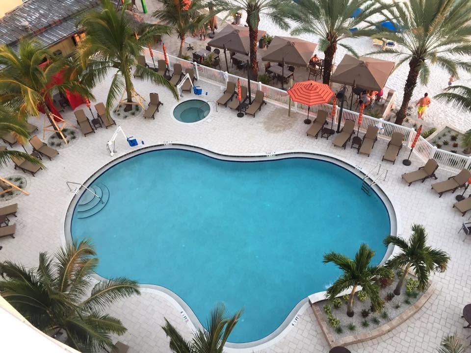 Aerial view of a resort pool with lounge chairs, palm trees, and thatched umbrellas.
