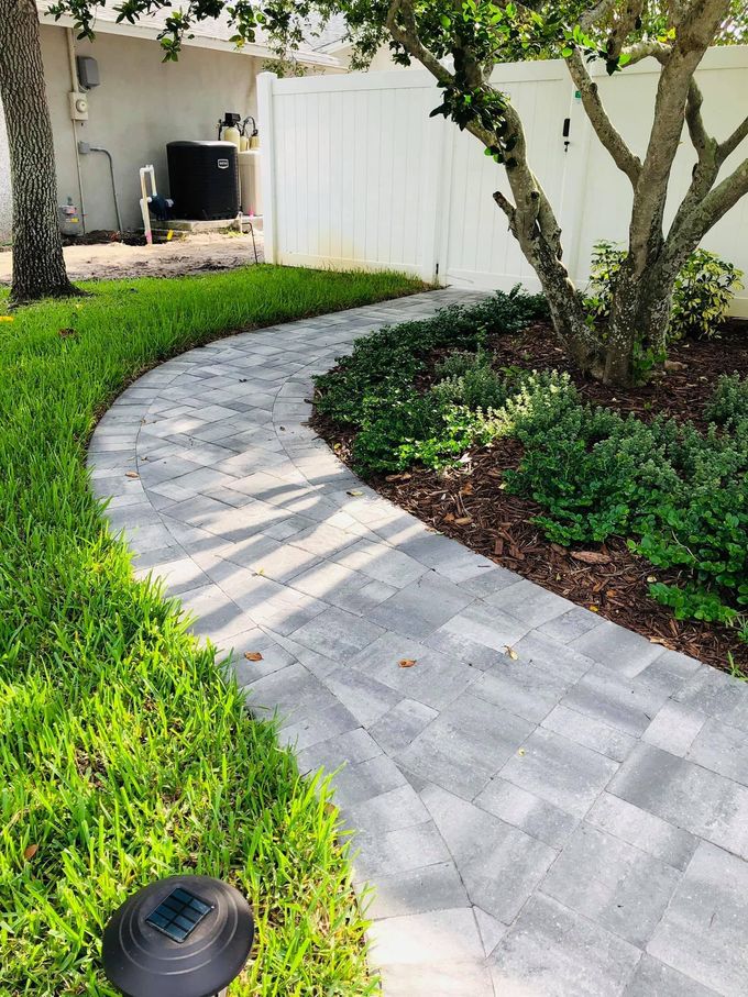 Curved brick paver walkway through a landscaped yard with grass, trees, and a white fence