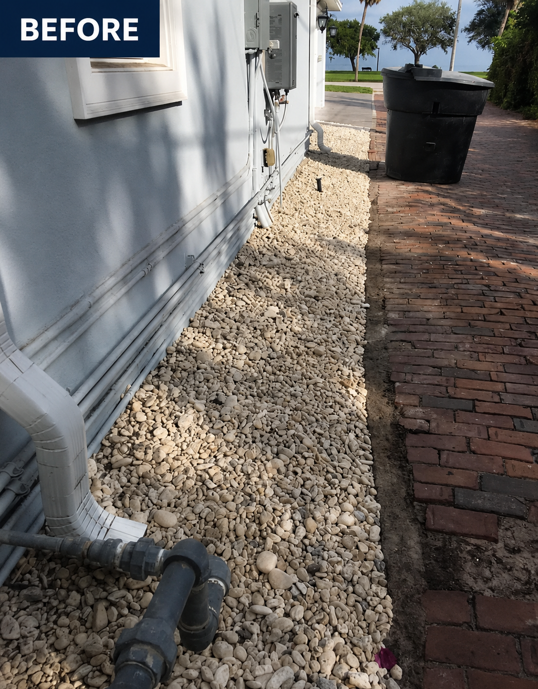 Before photo of a house side yard with white rocks beside a brick walkway and trash bins.