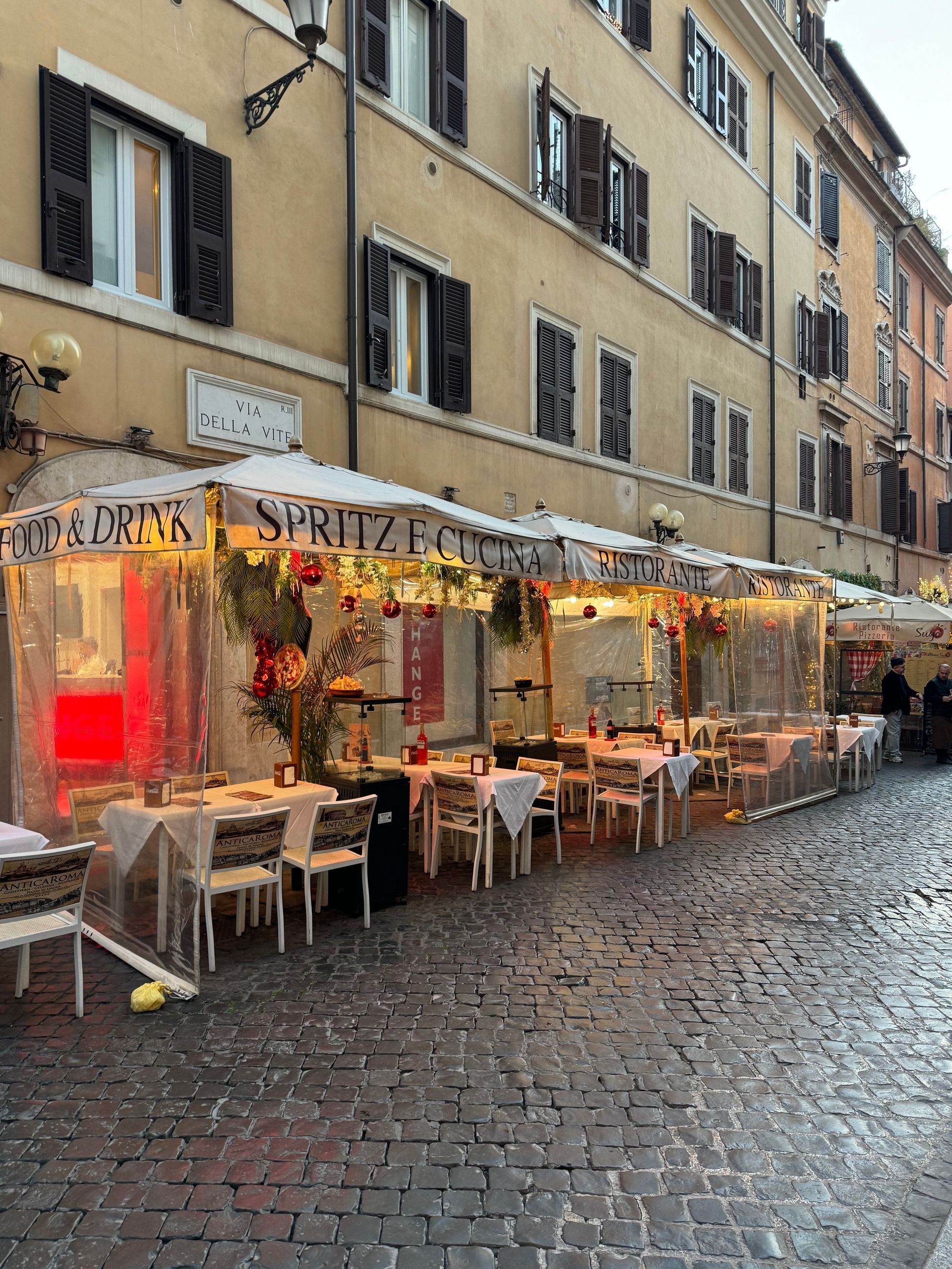 Rainy cobblestone street with a warmly lit outdoor café under beige buildings and red awnings