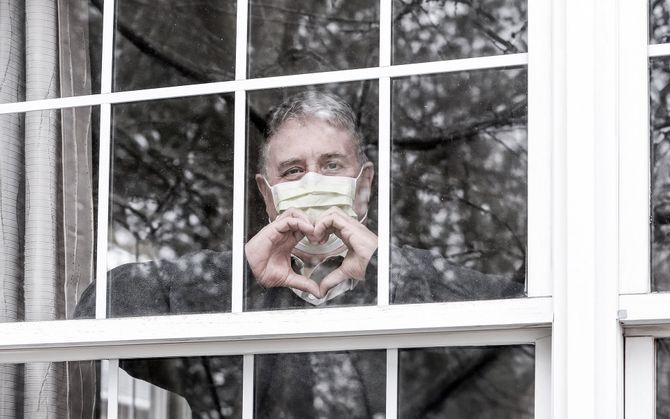 Man in mask looking out from window forming a heart with his hands
