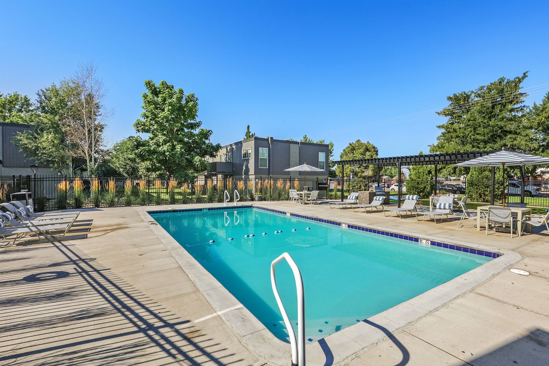 An outdoor swimming pool surrounded by a concrete deck, lounge chairs, and patio tables under a pergola.