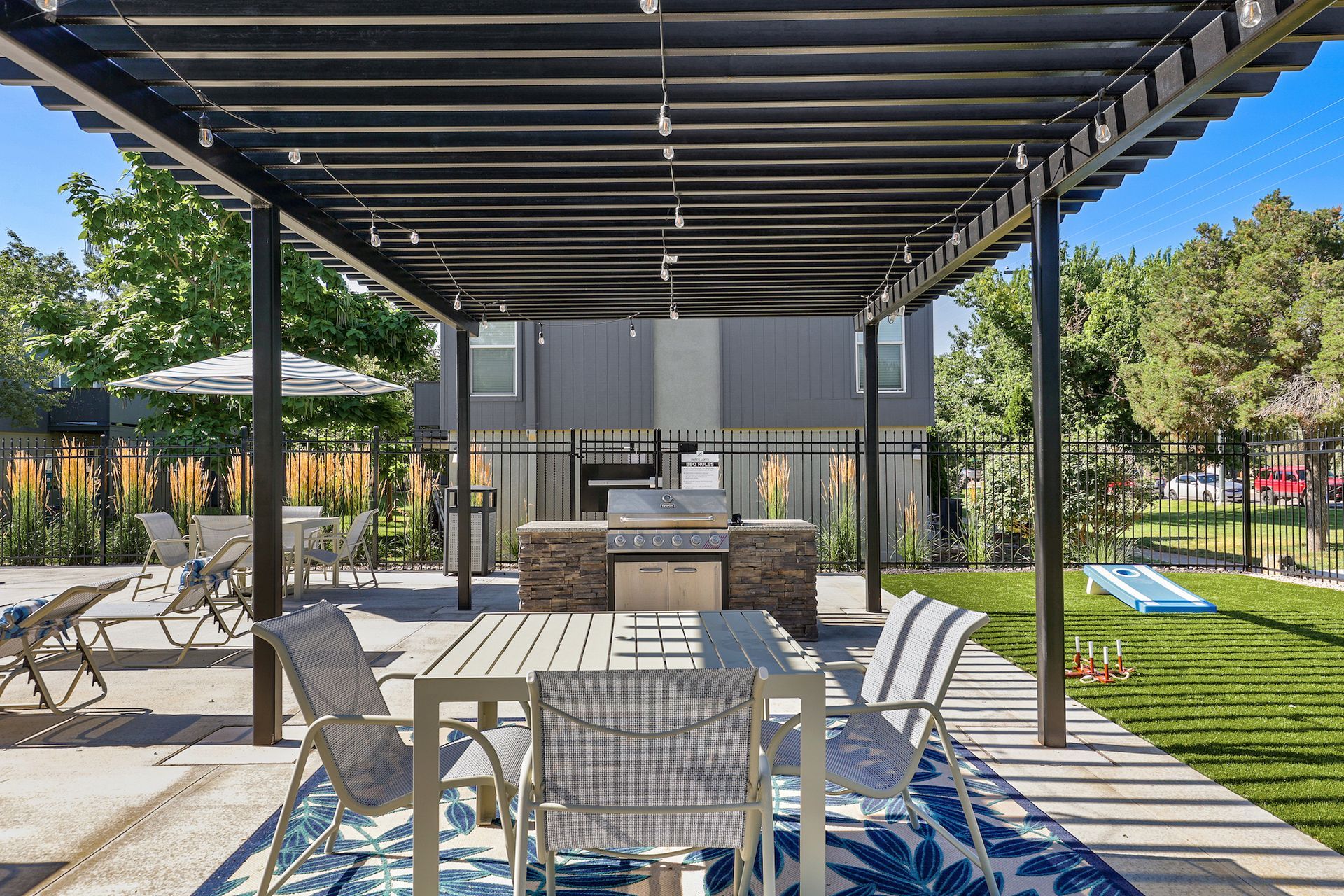 A shaded outdoor patio with a dining table, chairs, a built-in grill station, and a lawn with a small slide.