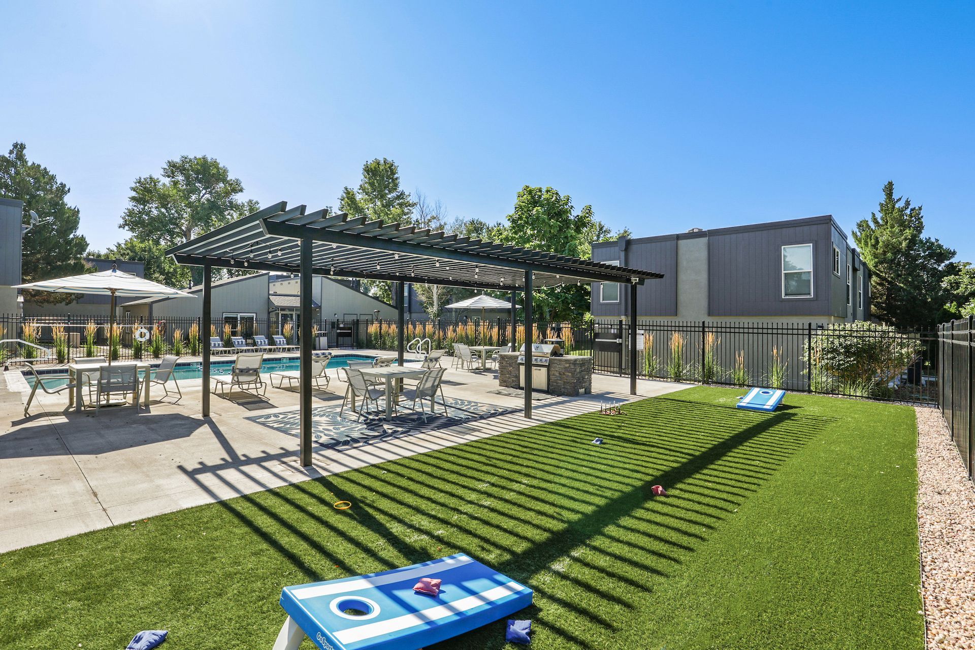 An apartment courtyard featuring a swimming pool, shaded pergola, outdoor furniture, and a cornhole set on a grassy lawn.