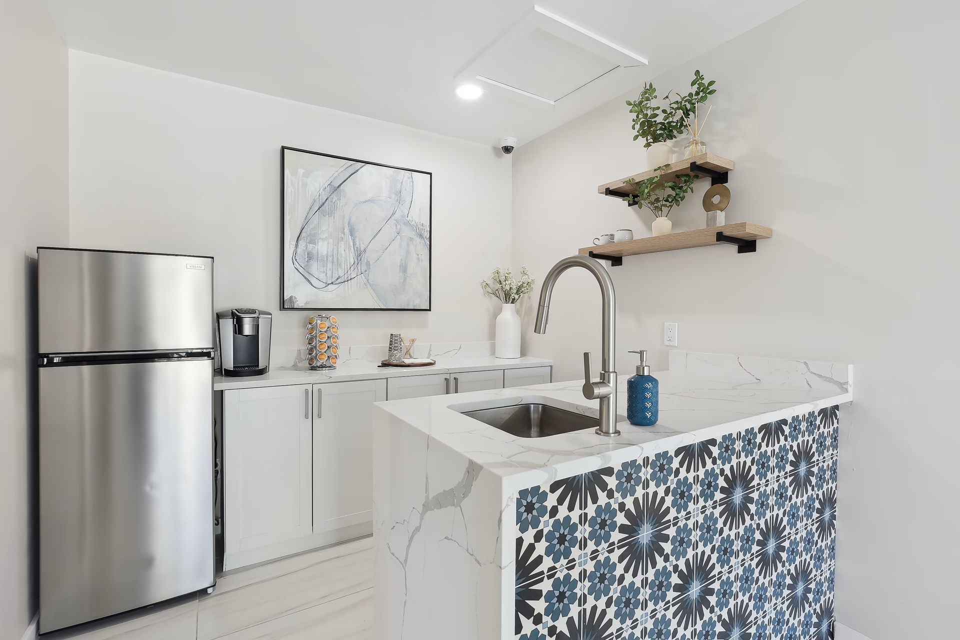 A small kitchenette with a stainless steel refrigerator, white cabinets, marble counters, and a blue patterned bar front.