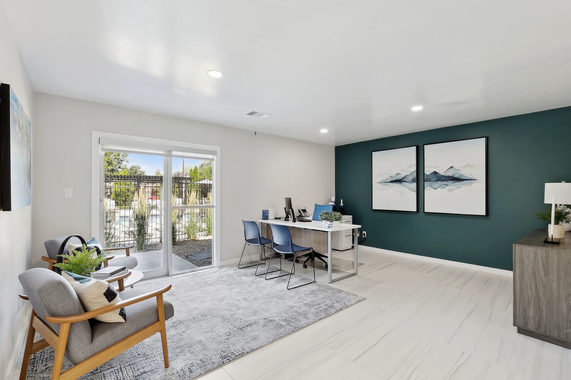 A modern office with a desk, two blue chairs, an armchair on a patterned rug, and wall art against a deep teal accent wall.