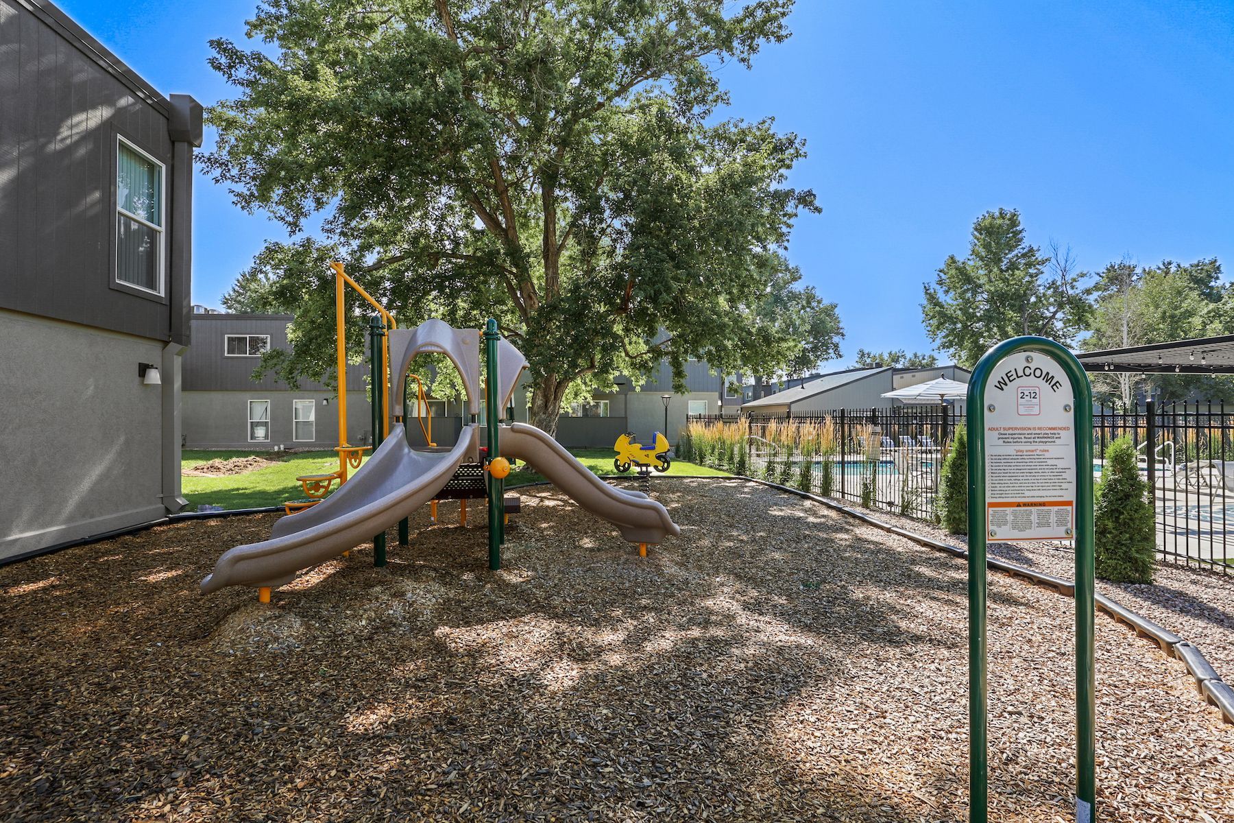 A playground with a brown plastic slide and wood chip ground surface, set next to an apartment building and pool area.