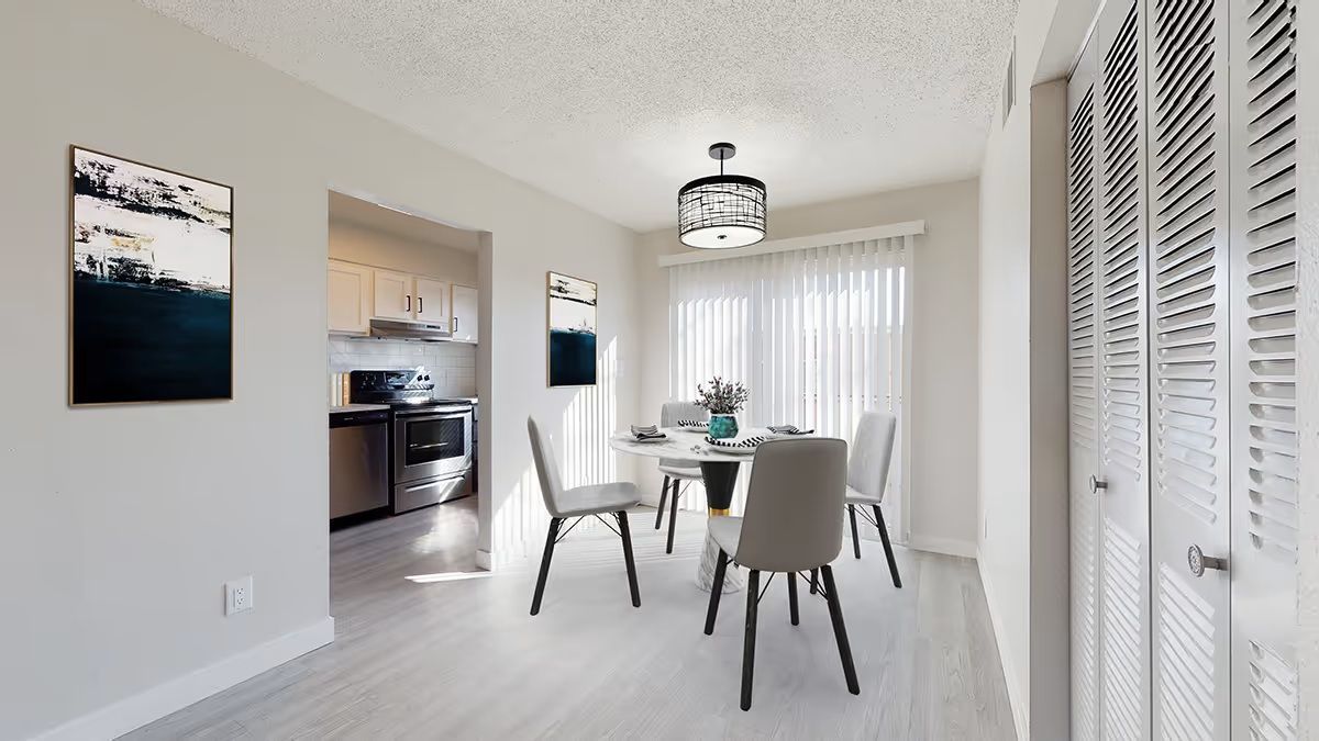 A modern dining area with a round table, four gray chairs, a pendant light, and an open entryway to a kitchen.
