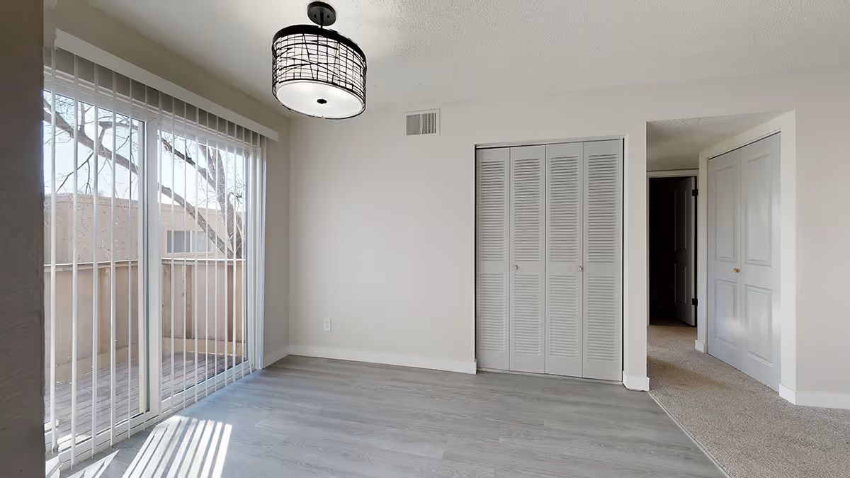A light-filled room with gray wood-look flooring, a modern chandelier, vertical blinds, and bifold closet doors.