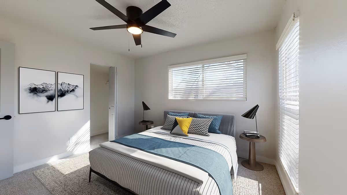A staged bedroom with a bed, two nightstands with lamps, and framed wall art, featuring a ceiling fan and natural light.