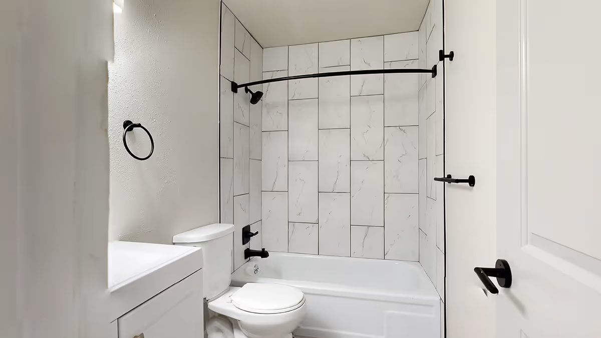 A small bathroom featuring a white vanity, toilet, and a bathtub with white marble-patterned wall tiles and black fixtures.